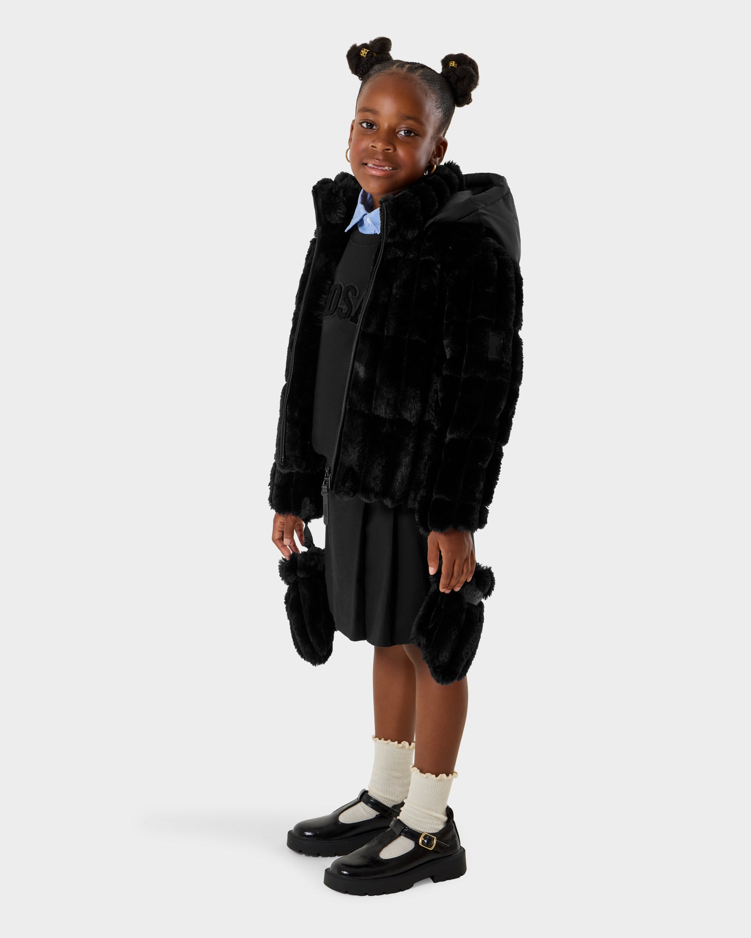 A young girl smiles against a white background, wearing the Rudsak ANASTASIA K - 0125000 Black faux fur shell coat with a black skirt, black shoes, white socks, and a blue-collared shirt. Her hair is styled in two buns.