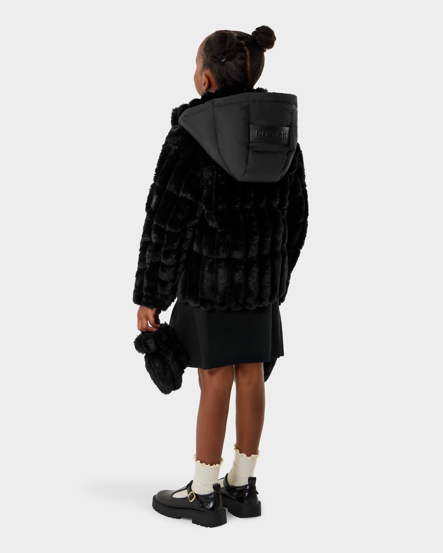 A young girl faces away, wearing the Rudsak ANASTASIA K - 0125000 Black faux fur hooded coat with a black skirt, white frilly socks, and black shoes. She holds a small black purse against a plain white background.