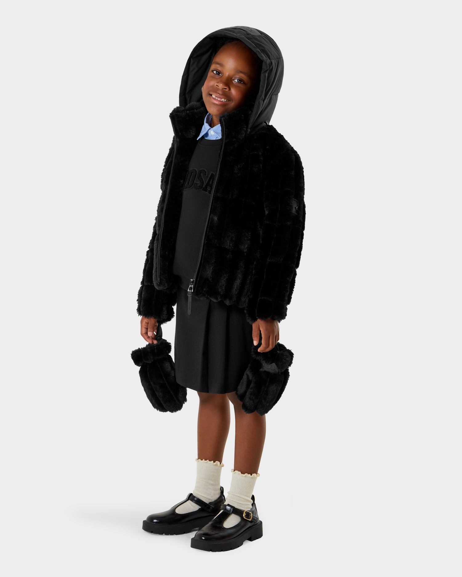 A young child wears the Rudsak ANASTASIA K - 0125000 Black faux fur shell jacket with hood, black skirt, white socks, and black shoes, smiling with matching furry accessories against a plain light background.