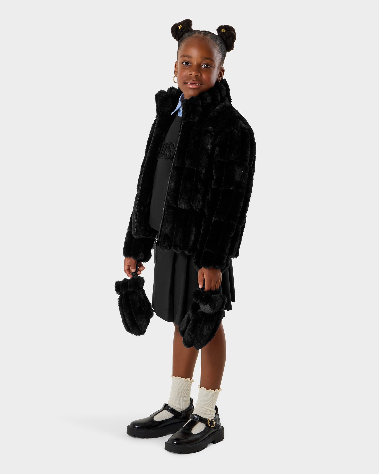 A young girl with two buns smiles, wearing Rudsak’s ANASTASIA K - 0125000 Black faux fur shell jacket and matching small black fur bag, paired with a black dress, frilled white socks, and black shoes.