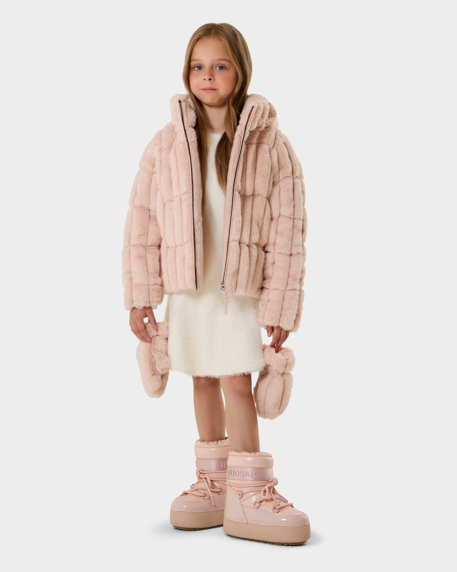 A young girl with long hair wears the Rudsak ANASTASIA K - 0125000 Pastel Pink faux fur shell jacket over a white dress with matching pink moon boots, holding the cuffs and standing against a plain light background.