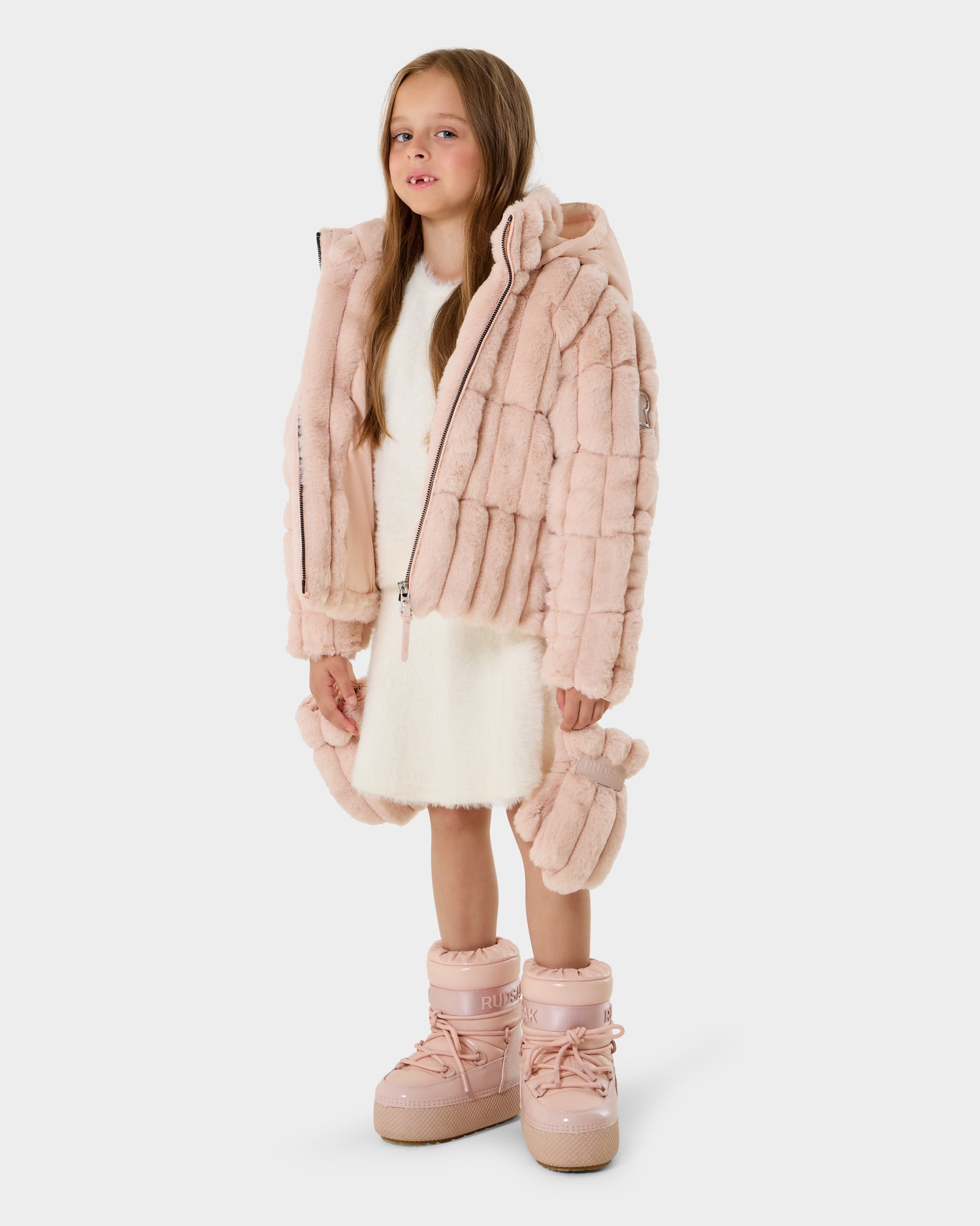 A young girl with long hair stands against a white background, wearing the Rudsak ANASTASIA K - 0125000 Pastel Pink plush hooded jacket, paired with a white dress and matching pink winter boots.