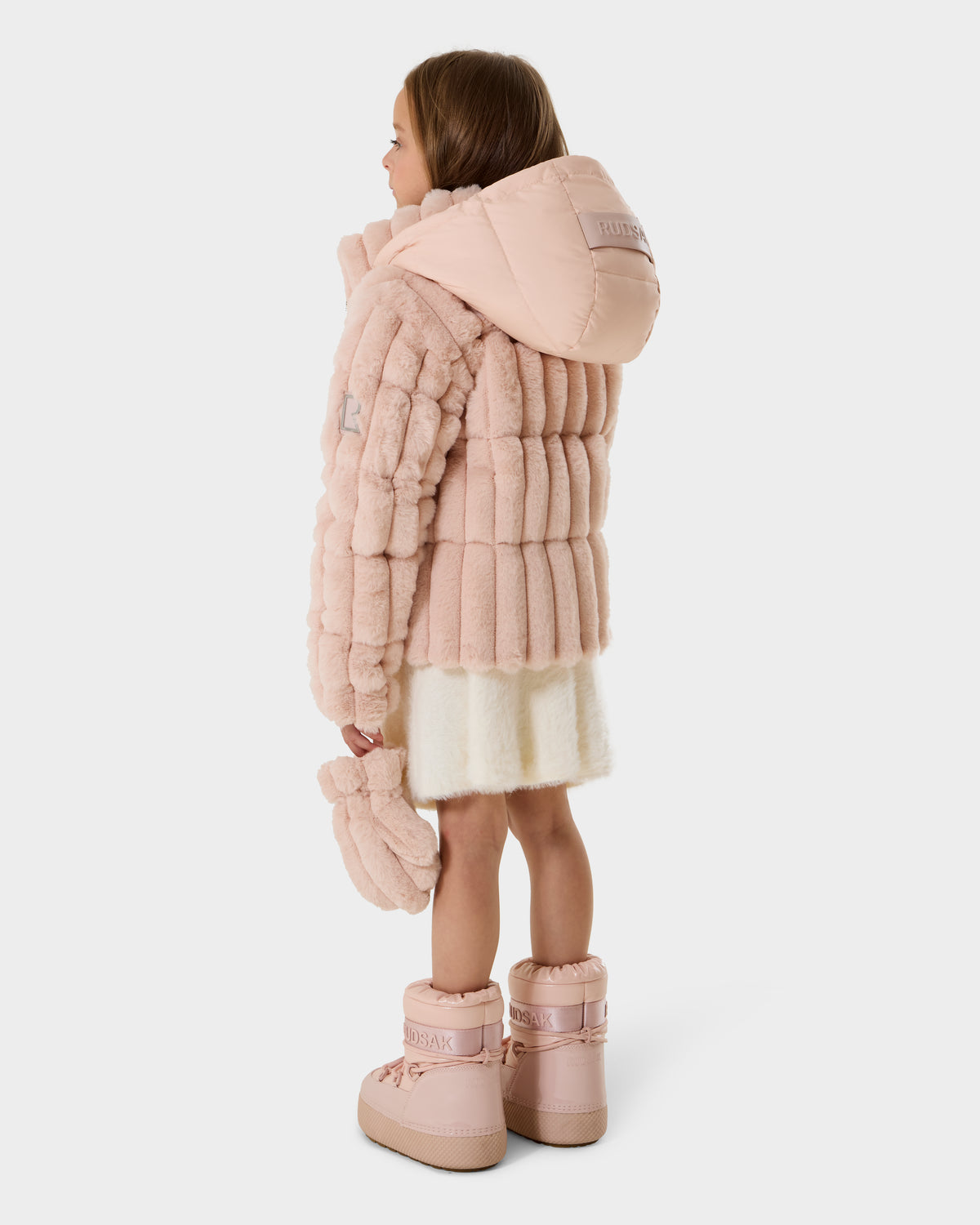 A young girl stands sideways in a Rudsak ANASTASIA K - 0125000 Pastel Pink faux fur shell jacket with hood, matching mitten in one hand, white skirt, and pastel pink winter boots against a plain white background.
