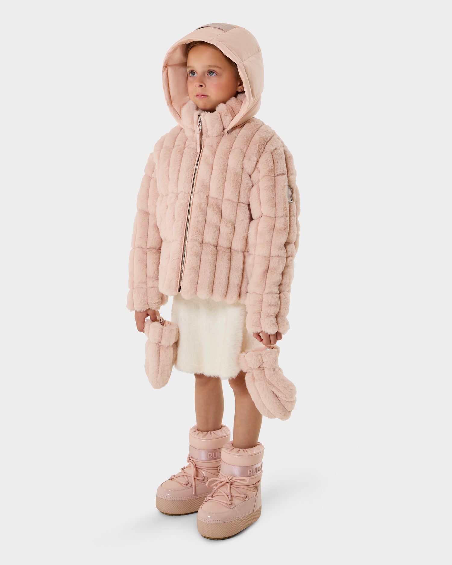 A young child stands in a Rudsak ANASTASIA K - 0125000 Pastel Pink hooded puffer jacket with matching mittens and pink boots, wearing a white skirt, looking straight ahead against a plain white background.