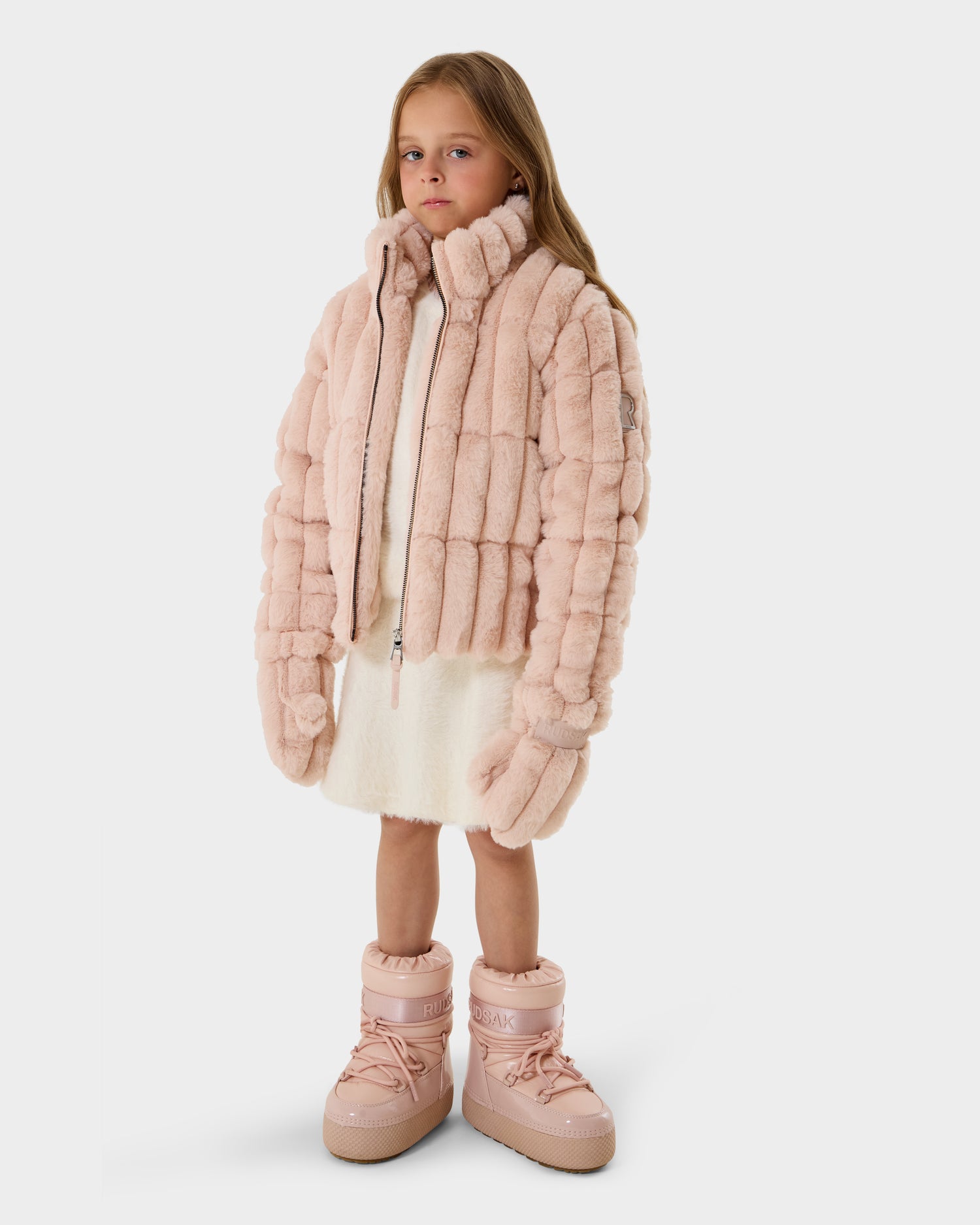 A young girl with long, straight hair and a neutral look stands against a plain white background, wearing the Rudsak ANASTASIA K - 0125000 Pastel Pink quilted jacket, a white dress, and chunky pale pink boots.