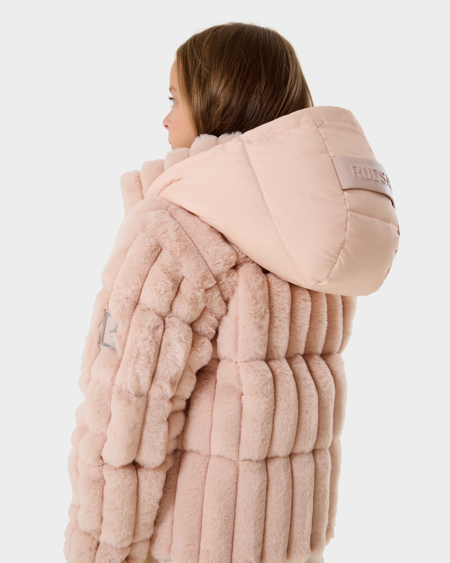 A person with light brown hair is seen from the back wearing the Rudsak ANASTASIA K - 0125000 Pastel Pink puffer jacket, featuring a textured finish, large hood, and gray sleeve patch against a plain white background.