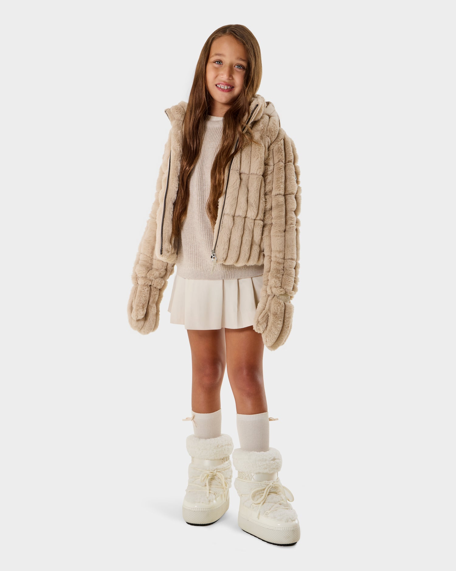 A young girl with long brown hair wears the Rudsak ANASTASIA K - 0125000 Vanilla puffer, paired with a white sweater, cream skirt, knee-high socks, and chunky white boots, smiling against a plain white background.