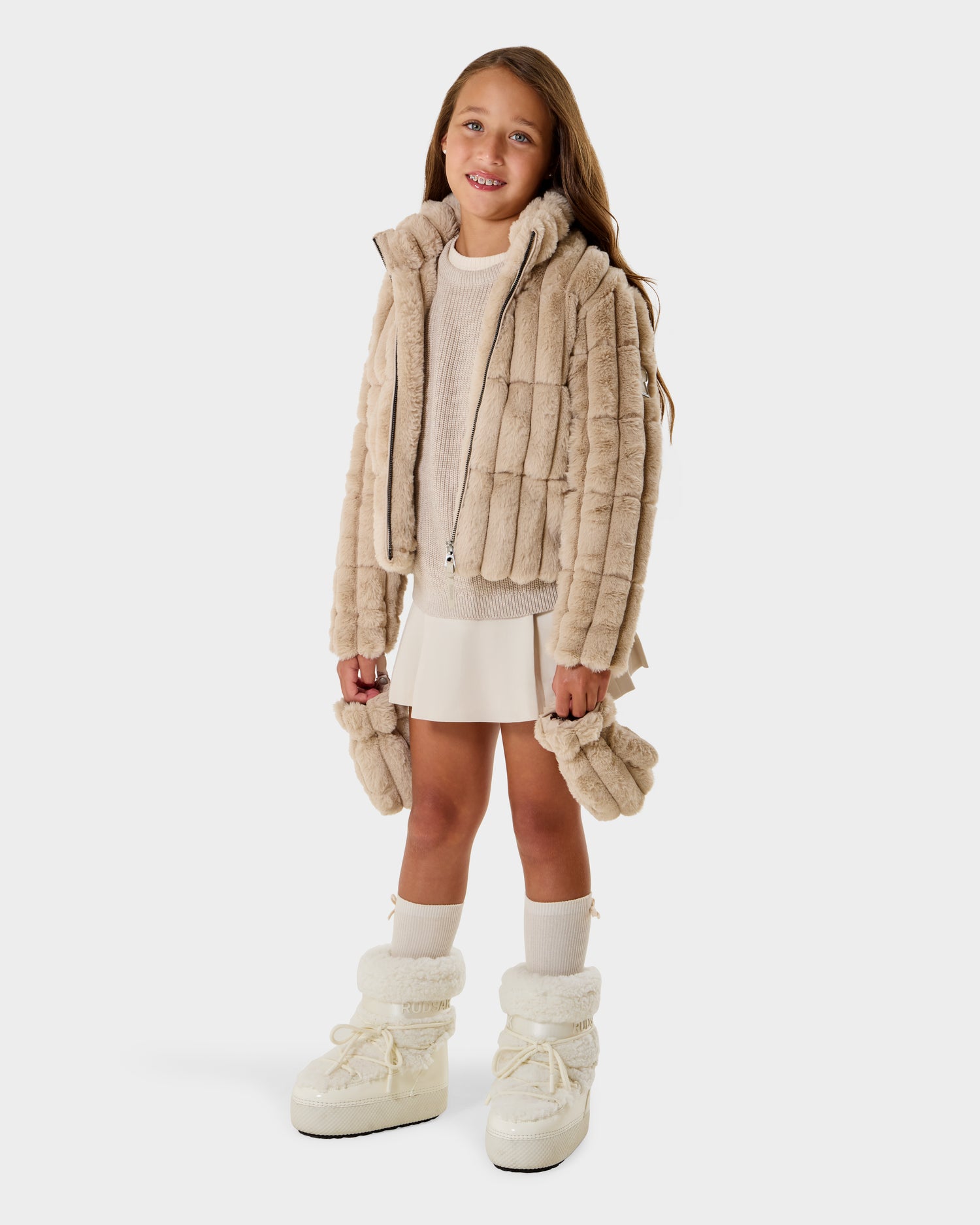 A smiling girl wears the Rudsak ANASTASIA K - 0125000 Vanilla faux fur shell jacket with matching mittens, white sweater, skirt, knee-high socks, and winter boots, standing on a white background.