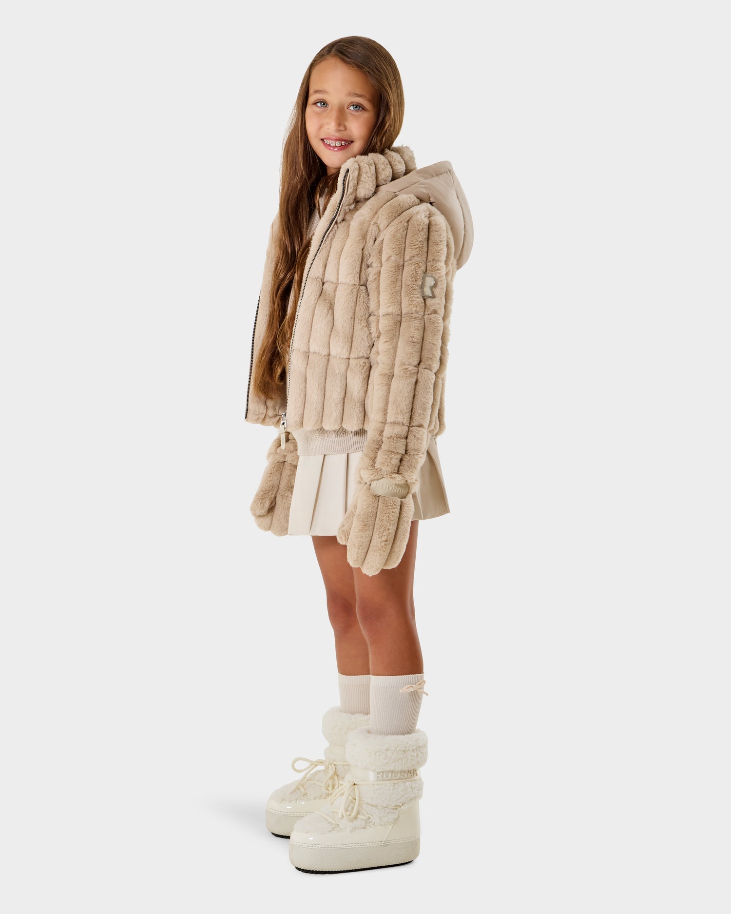 A young girl wears the ANASTASIA K - 0125000 Vanilla shell jacket by Rudsak, paired with mittens, a pleated skirt, knee-high socks, and white boots, smiling as she stands against a plain white background.