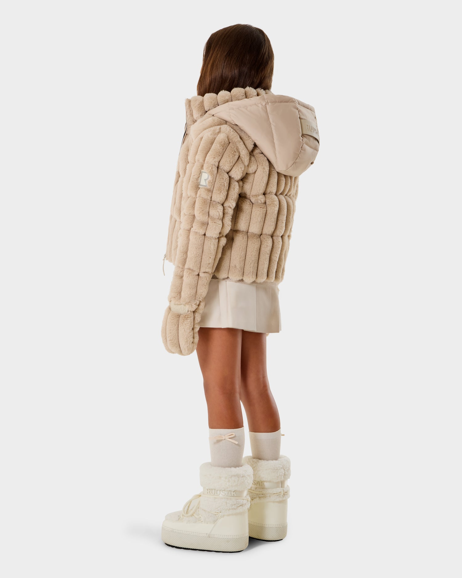 A young girl with long brown hair faces away, wearing the Rudsak ANASTASIA K - 0125000 Vanilla puffer jacket, a cream skirt, white socks, and chunky white boots against a plain background.