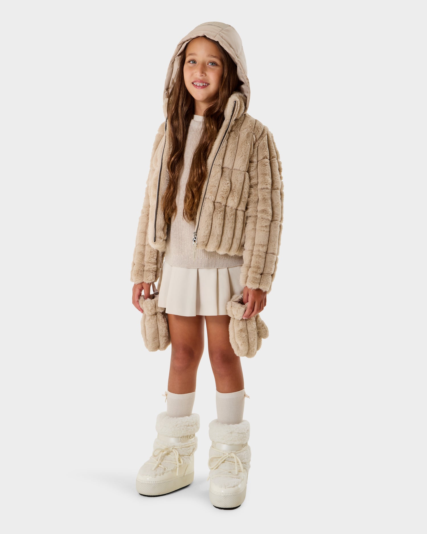 A young girl with long brown hair smiles in the Rudsak ANASTASIA K - 0125000 Vanilla beige faux fur shell jacket with hood, paired with a cream sweater, pleated skirt, white socks, and chunky white boots against a plain white background.