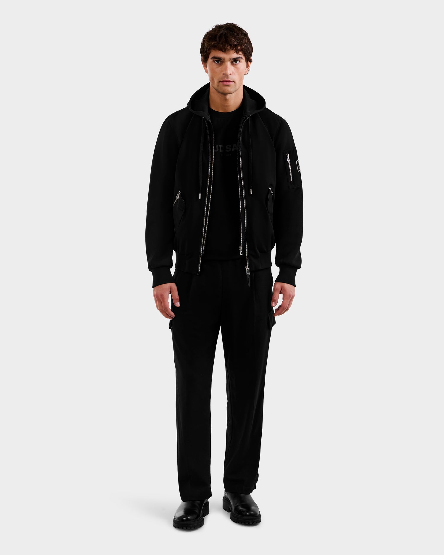 A man stands facing forward against a white background, wearing the Rudsak ANDRE - 6124020 Black bomber jacket with removable hooded bib, paired with black t-shirt, pants, and shoes. His arms are relaxed by his sides.