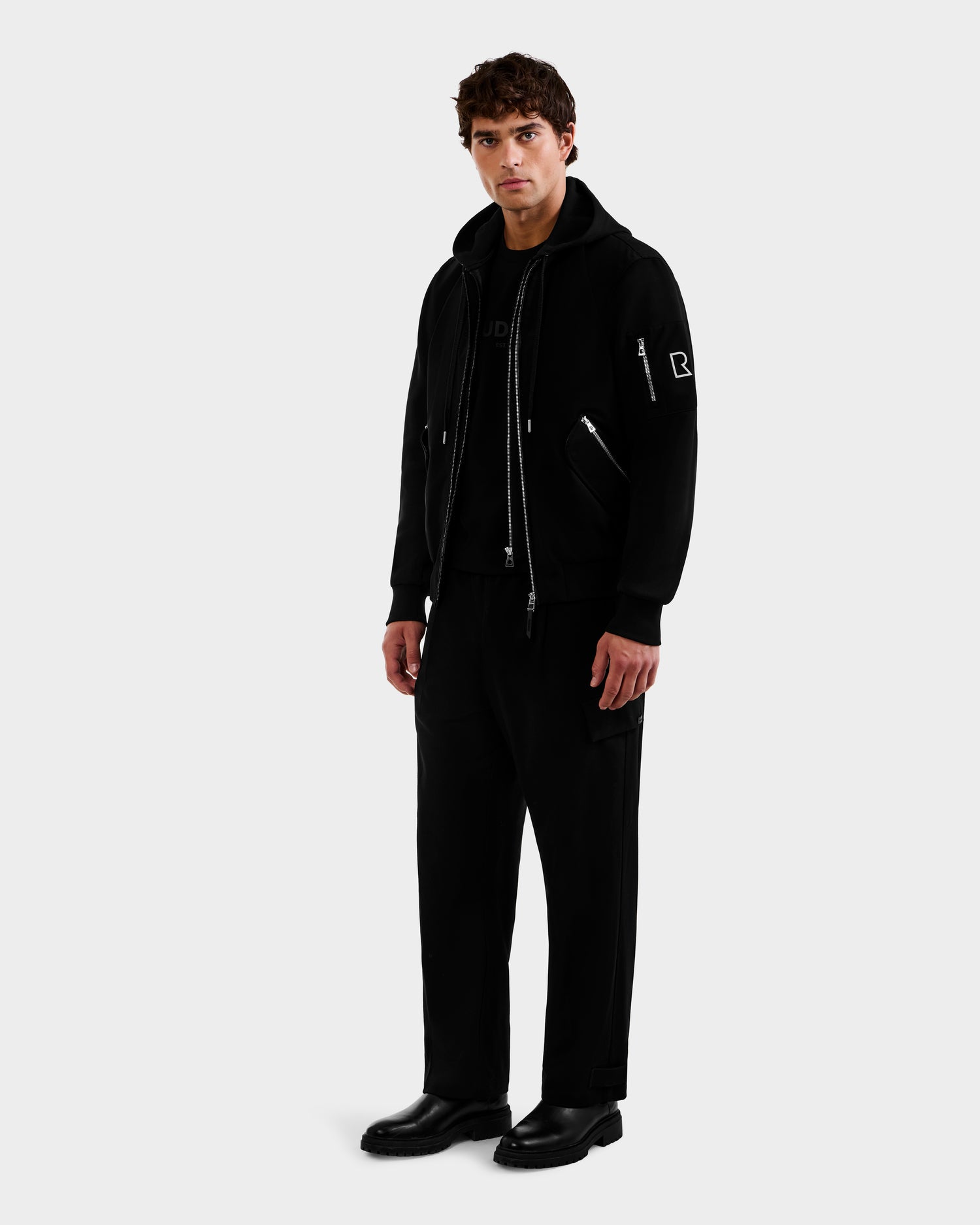 A man with short, curly brown hair stands facing forward against a plain white background, wearing the Rudsak ANDRE - 6124020 Black men’s lightweight bomber jacket with removable hooded bib, black pants, and black shoes. His hands rest by his sides.
