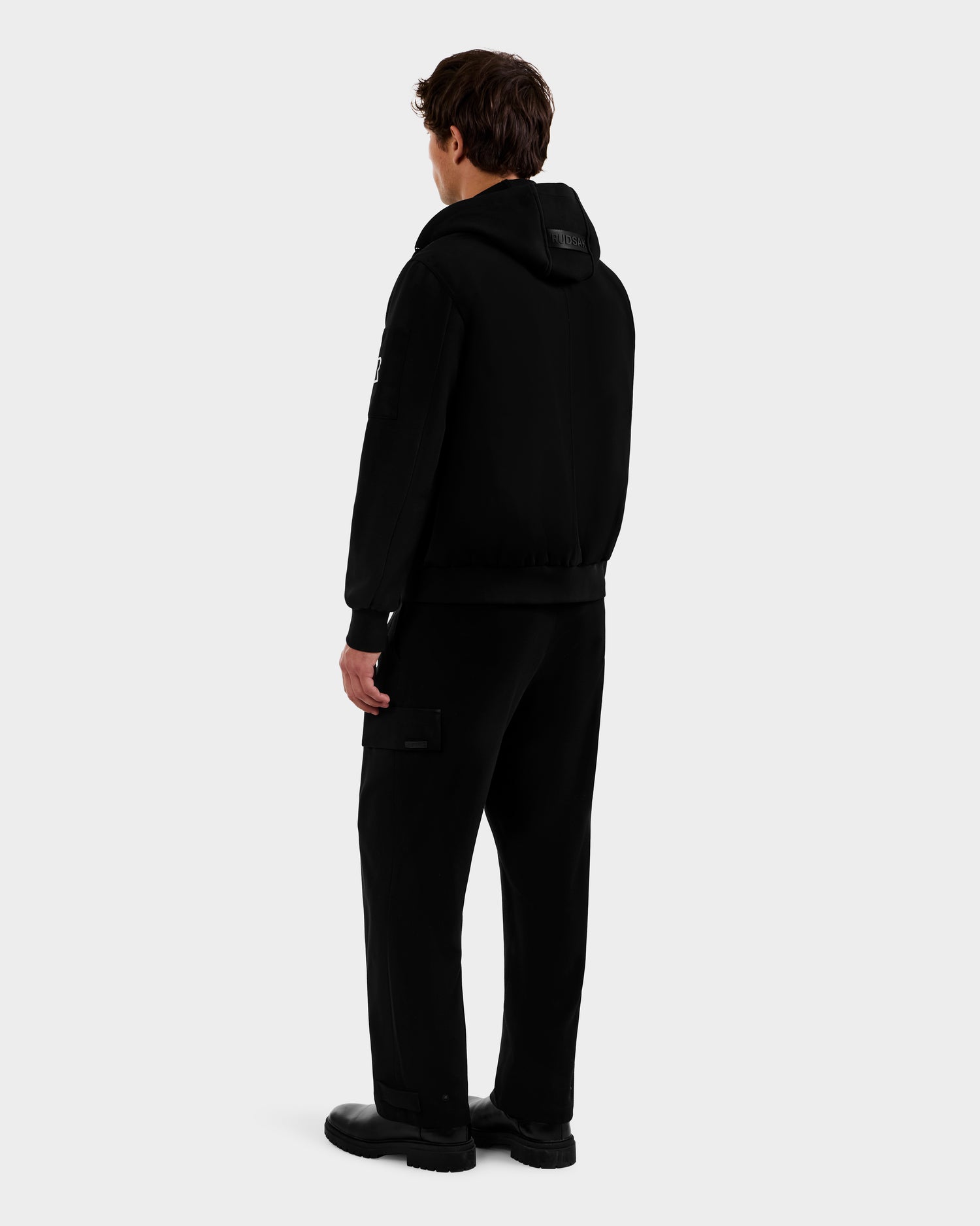 A person with short brown hair, facing away, models Rudsak's ANDRE - 6124020 Black men’s lightweight bomber jacket with removable hooded bib, paired with black pants and shoes against a plain light gray background.