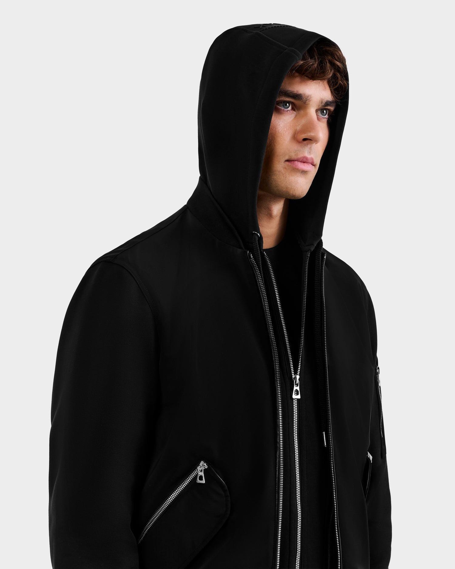 A young man wearing the Rudsak ANDRE - 6124020 Black men's lightweight bomber jacket with multiple zippers stands against a plain light background, looking slightly to the side with a neutral expression.