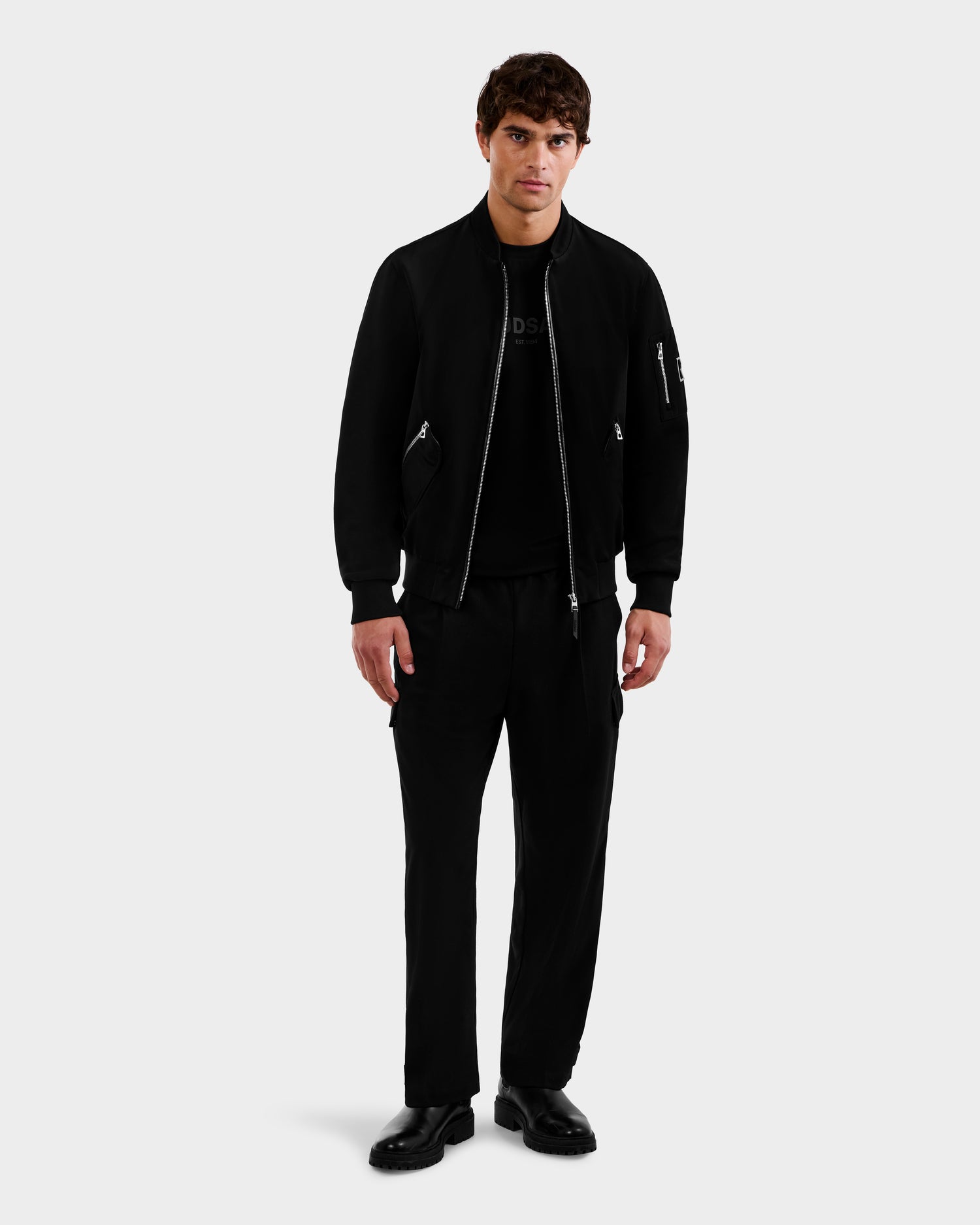 A man faces forward, wearing Rudsak’s ANDRE - 6124020 Black lightweight bomber jacket with removable hooded bib, paired with black pants and shoes, against a light gray background for a modern look.