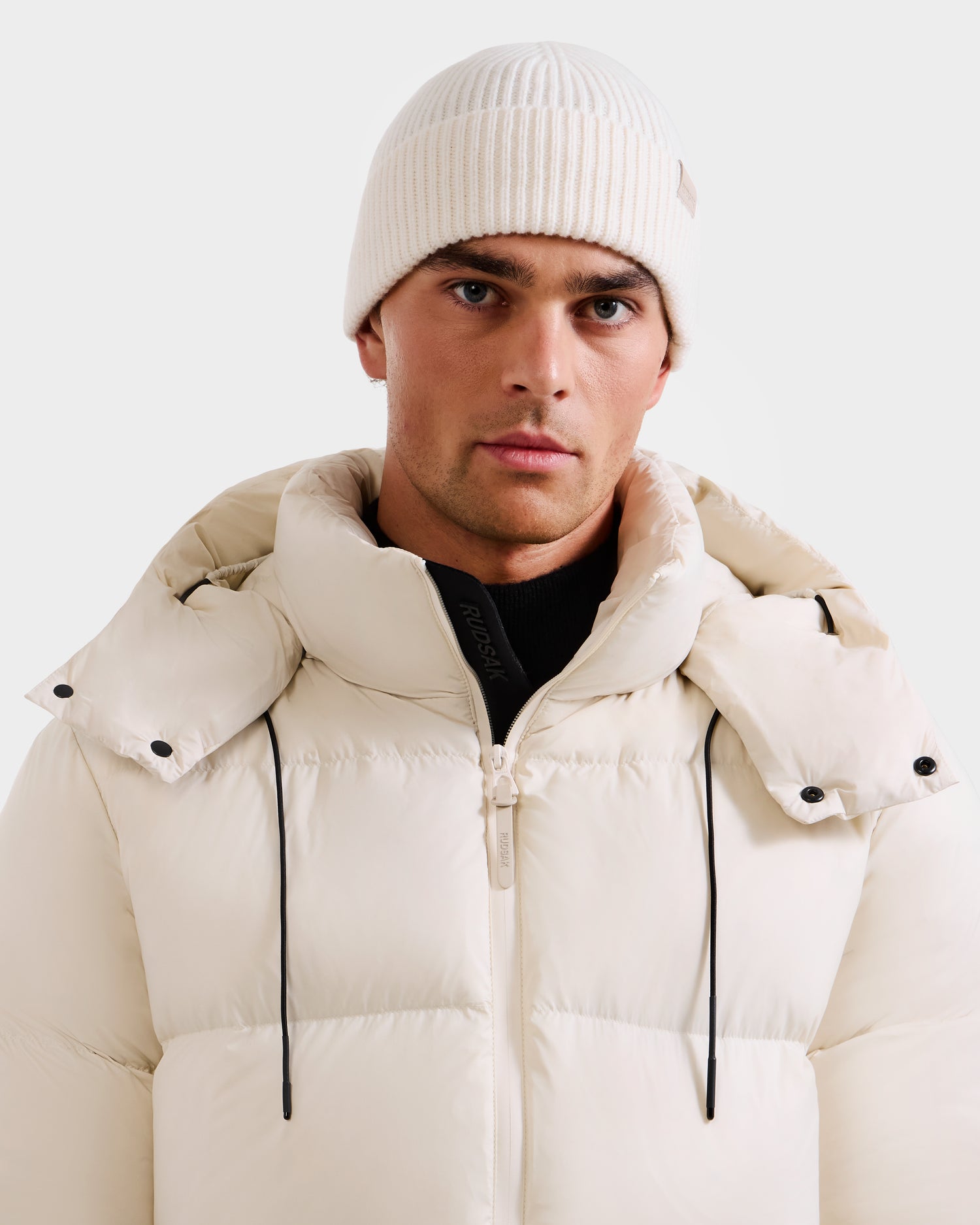 A man stands against a plain light background, looking directly at the camera in Rudsak’s ANDREY - U425507 Vanilla white puffer jacket with high collar and a unisex rib-knit beanie—an effortlessly stylish cold-weather essential.