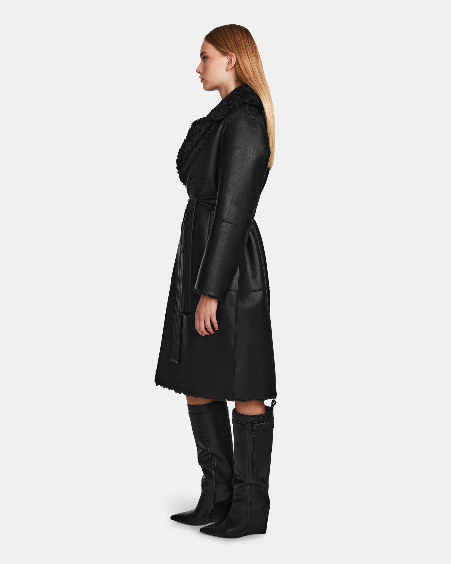 A woman with straight blonde hair poses in profile against a white background, wearing Rudsak’s ANNABELLE - L124544 Black vegan leather coat with a wide furry collar, waist belt, and knee-high black boots.