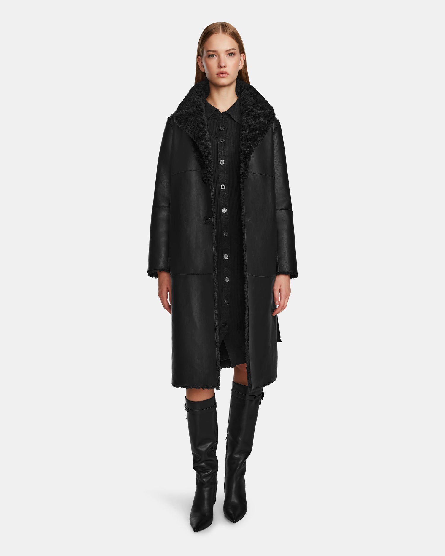 A woman faces forward wearing the ANNABELLE - L124544 Black shearling coat by Rudsak over a black buttoned dress and black knee-high boots, standing against a plain white background.