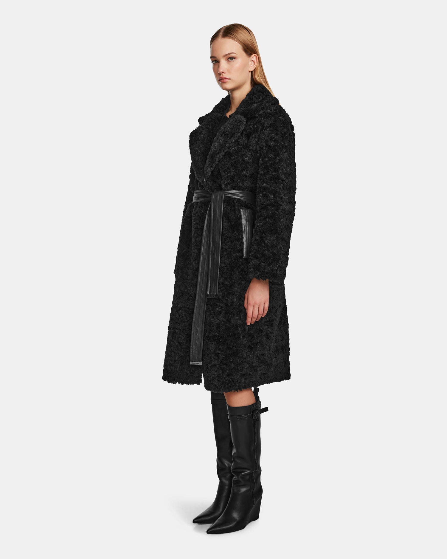A woman models the Rudsak ANNABELLE - L124544 Black textured vegan leather coat with a matching black belt and knee-high boots, standing slightly sideways against a plain white background.