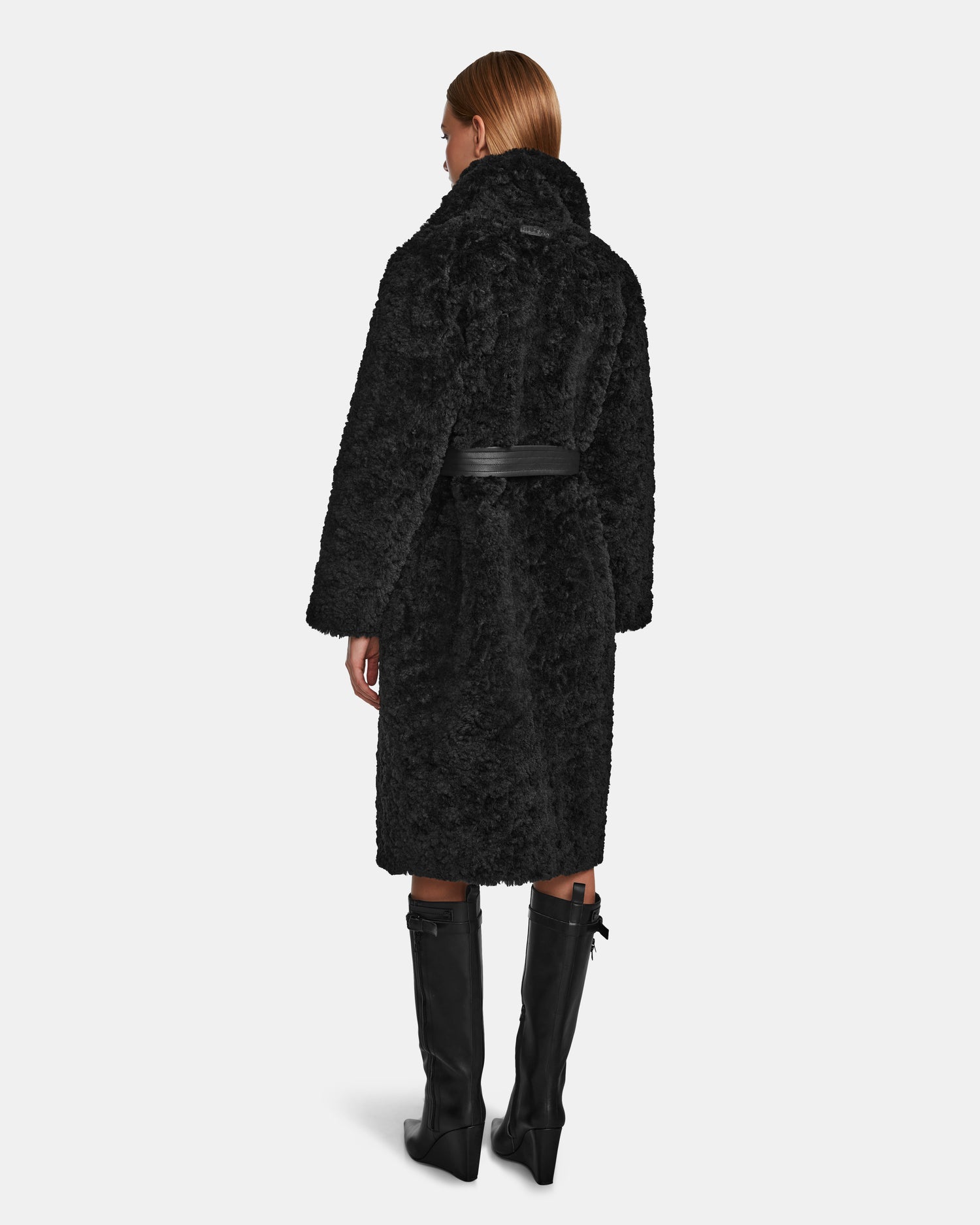 A woman with straight, light brown hair is seen from behind wearing the Rudsak ANNABELLE - L124544 Black knee-length coat with a black belt and tall black boots.