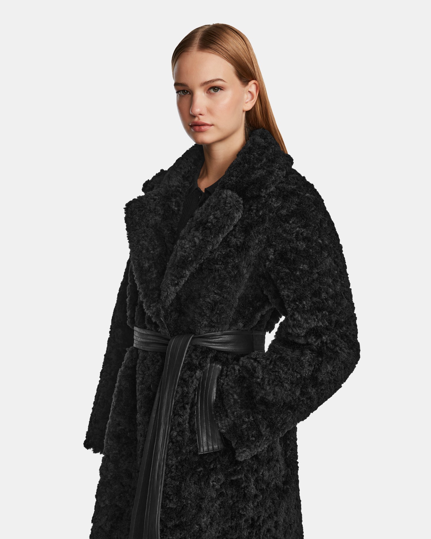 A woman with straight, light brown hair wears the Rudsak ANNABELLE - L124544 Black faux fur coat with a wide collar and black belt, standing against a plain light background.