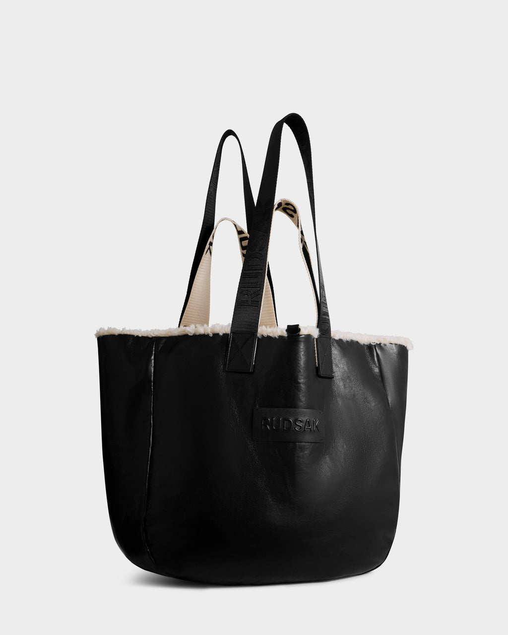 The Rudsak ANNIAH - 8325541 in Black/Vanilla is a black recycled leather tote with black handles, white faux shearling trim on the top edge, and an embossed RUDSAK logo on the front, shown against a plain light background.