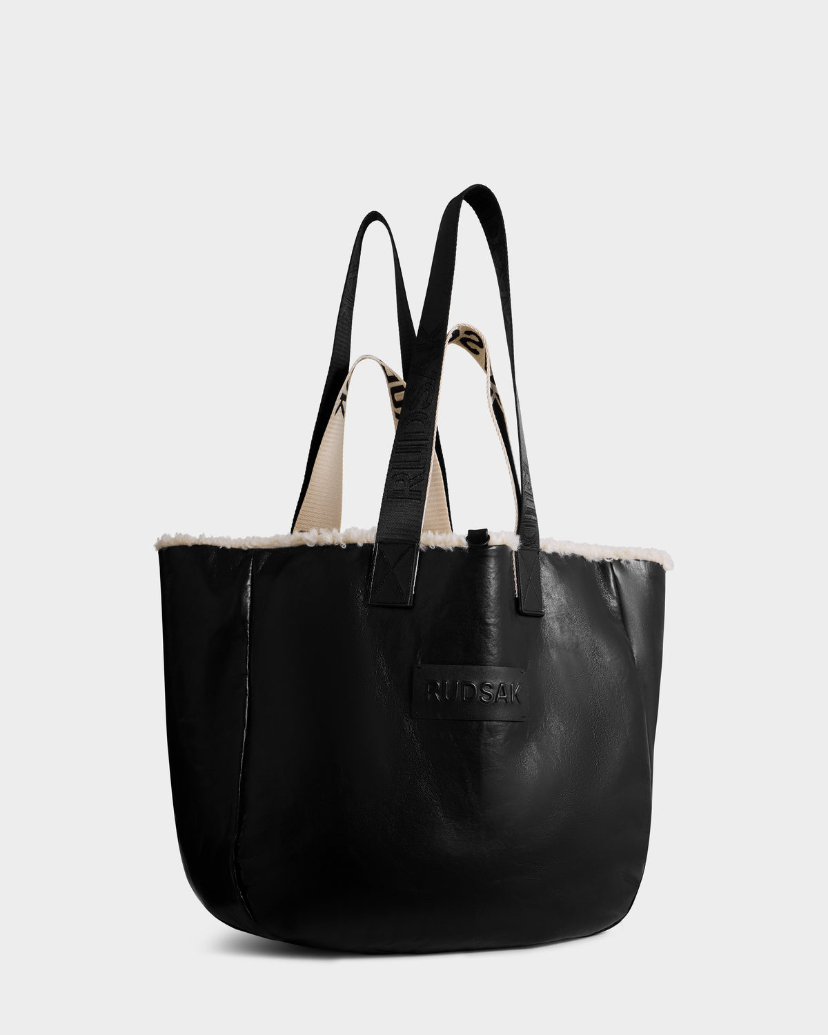 The Rudsak ANNIAH - 8325541 in Black/Vanilla is a black recycled leather tote with black handles, white faux shearling trim on the top edge, and an embossed RUDSAK logo on the front, shown against a plain light background.