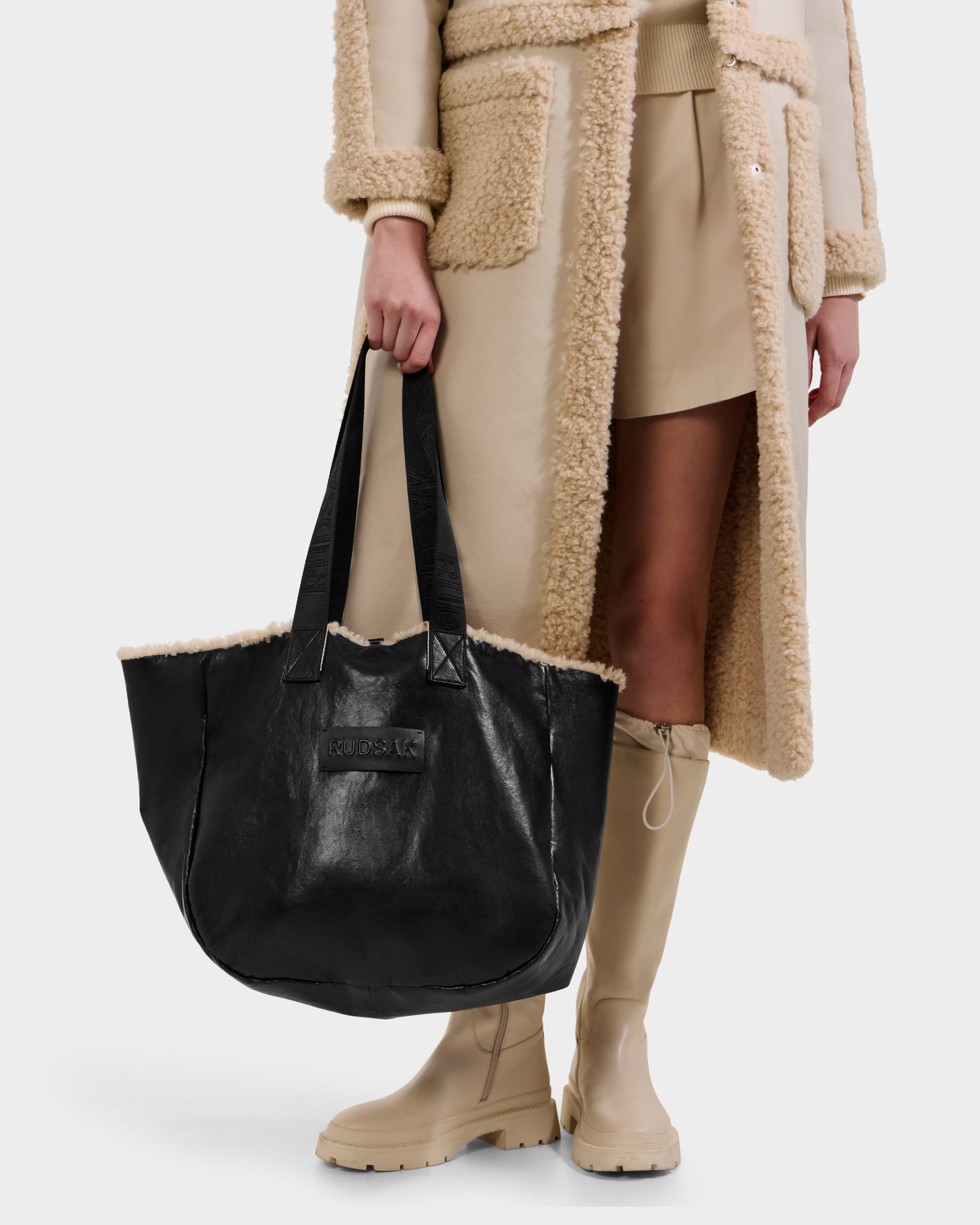 A person in a beige shearling coat, tan skirt, and beige knee-high boots holds the Rudsak ANNIAH - 8325541 Black faux shearling bag with black handles. Photo is cropped at the shoulders.