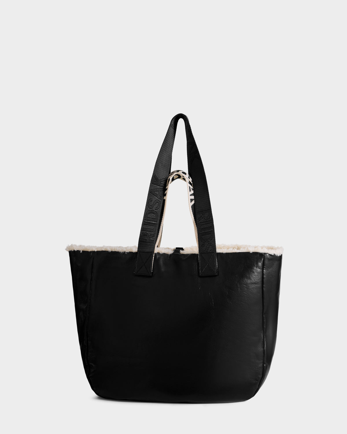 The Rudsak ANNIAH - 8325541 Black/Vanilla is a large reversible tote bag featuring a soft black faux shearling exterior, wide handles, and a fluffy white lining visible at the top, displayed against a light gray background.