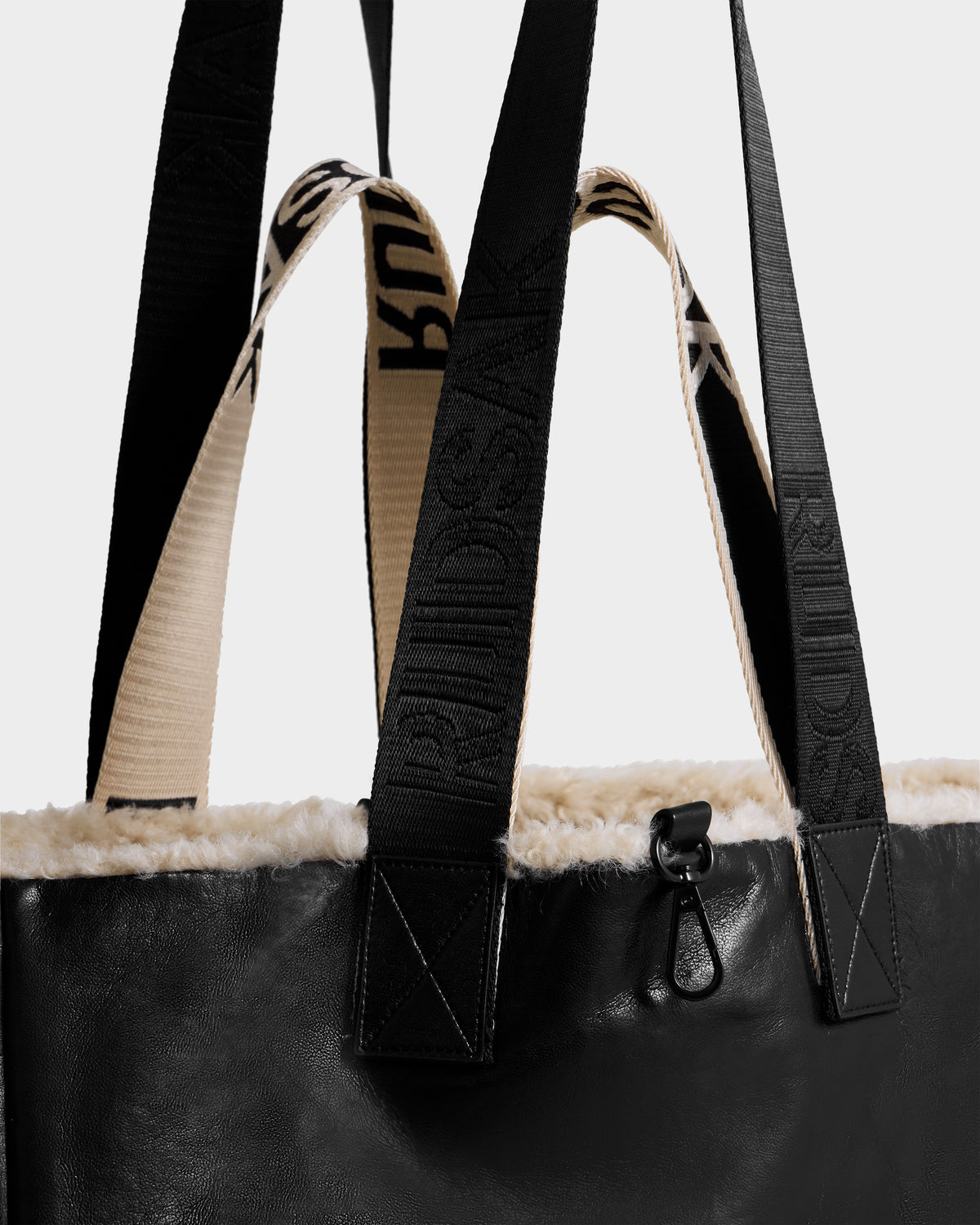 A close-up of the Rudsak ANNIAH - 8325541 tote in black/vanilla recycled leather with cream faux shearling trim, dual handles (one black, one cream with RUUDSAK text), and a black metal clip at the top edge.