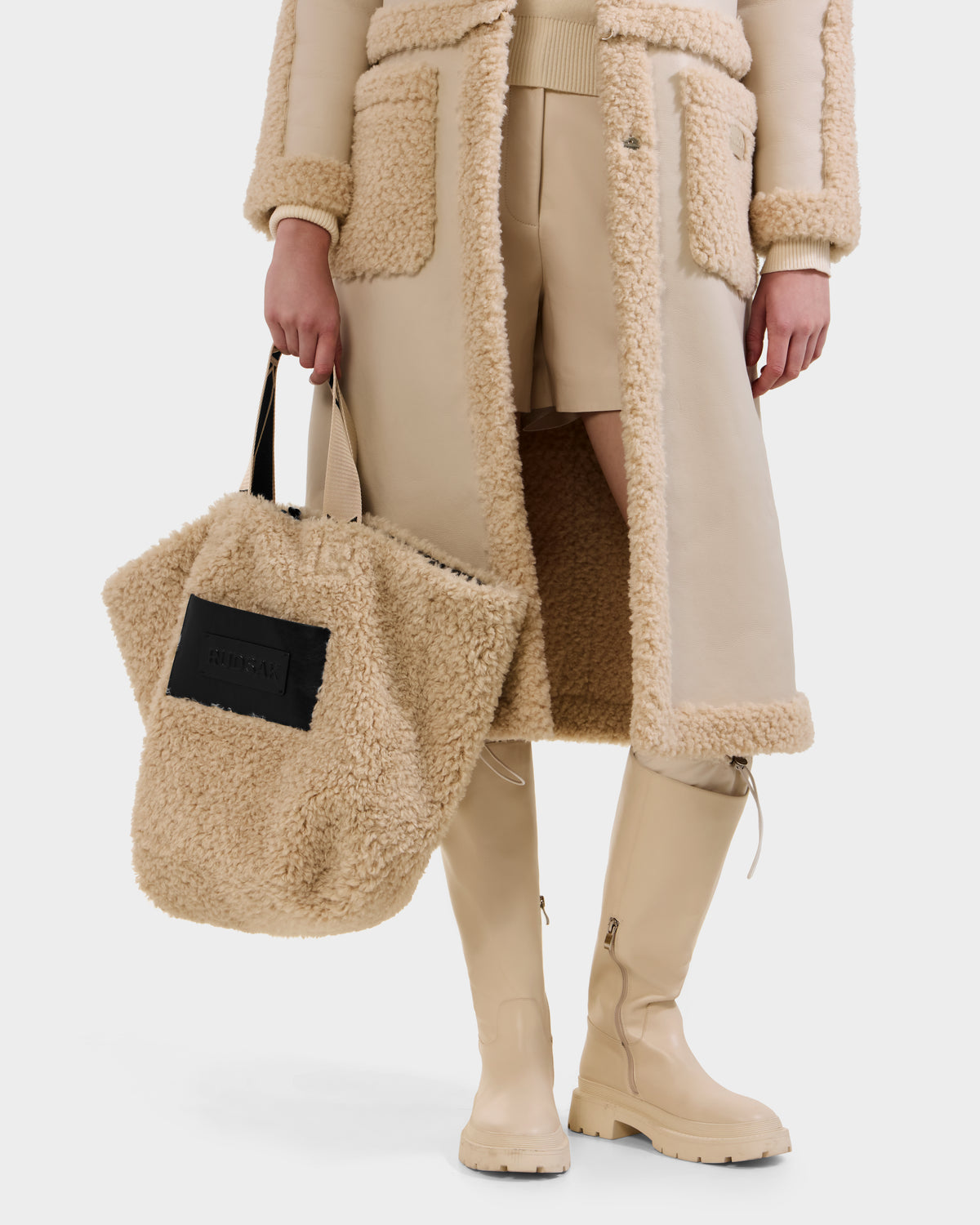 A person wears a beige faux shearling coat, matching shorts, and knee-high beige boots, holding Rudsak’s ANNIAH - 8325541 Black/Vanilla reversible tote in textured beige recycled leather with a black patch. Only the outfit and bag are visible; the face is not shown.