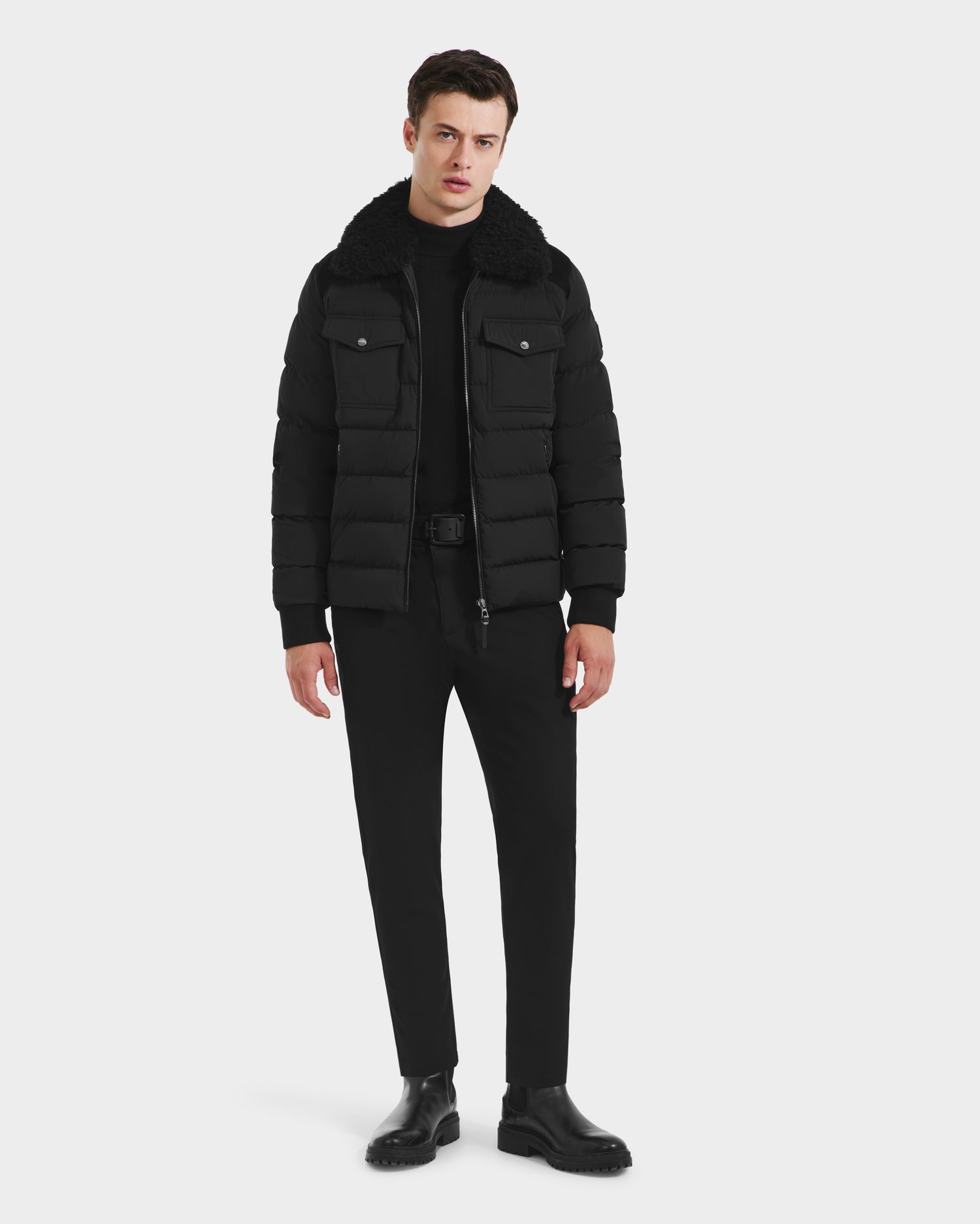 A man stands against a white background wearing the Rudsak ANTHONY - 6125042 Black quilted jacket with a shearling collar, paired with black pants, black boots, and a black shirt.
