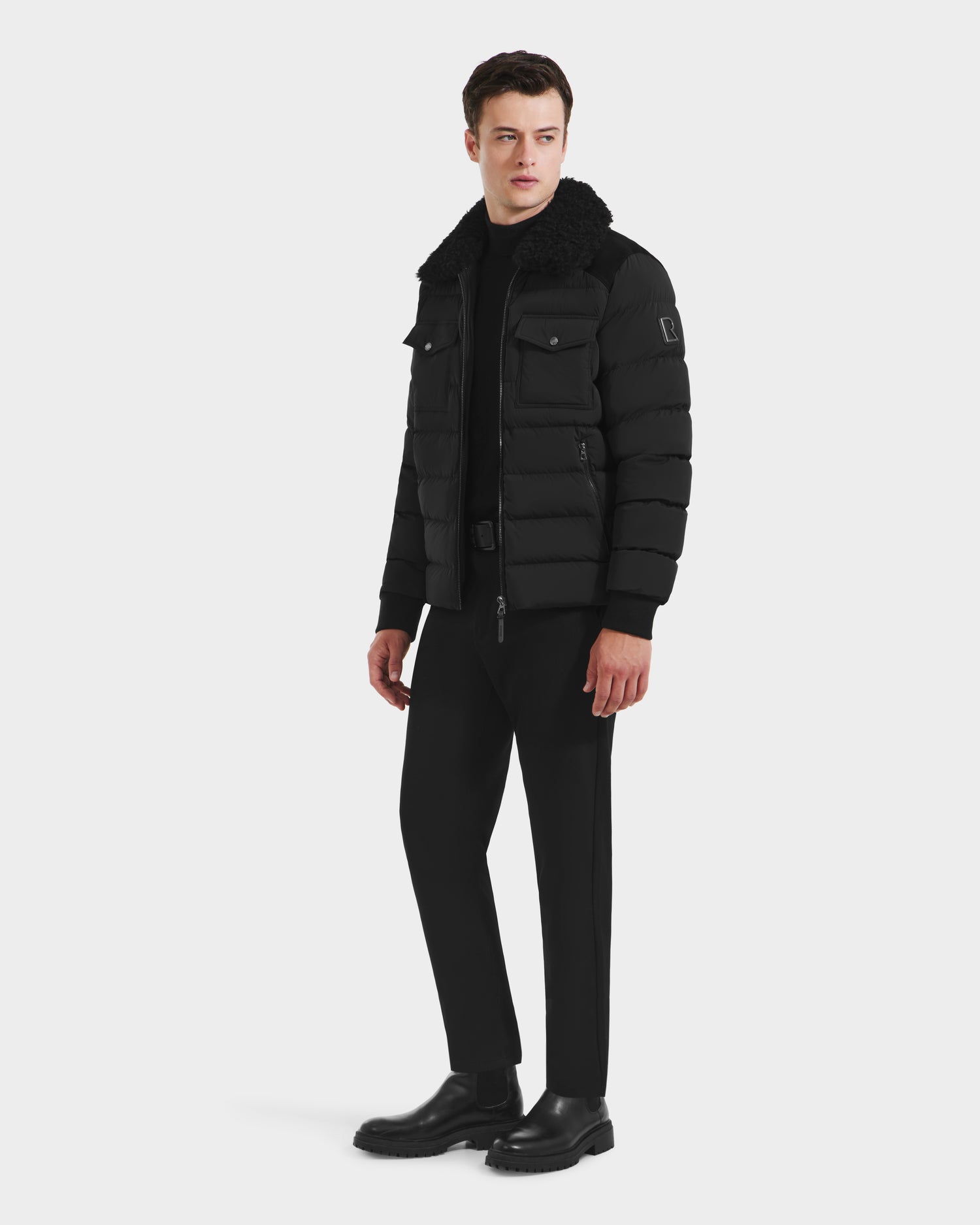 A man stands facing forward, wearing the Rudsak ANTHONY - 6125042 Black quilted jacket with shearling collar, paired with black pants and shoes against a plain white background.