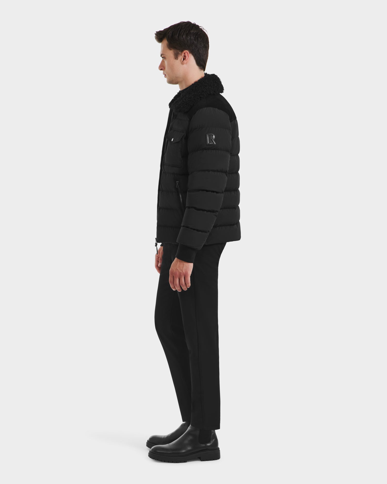 A man stands in profile wearing the Rudsak ANTHONY - 6125042 Black aviator jacket with a shearling collar, paired with black pants and boots, set against a plain white background.
