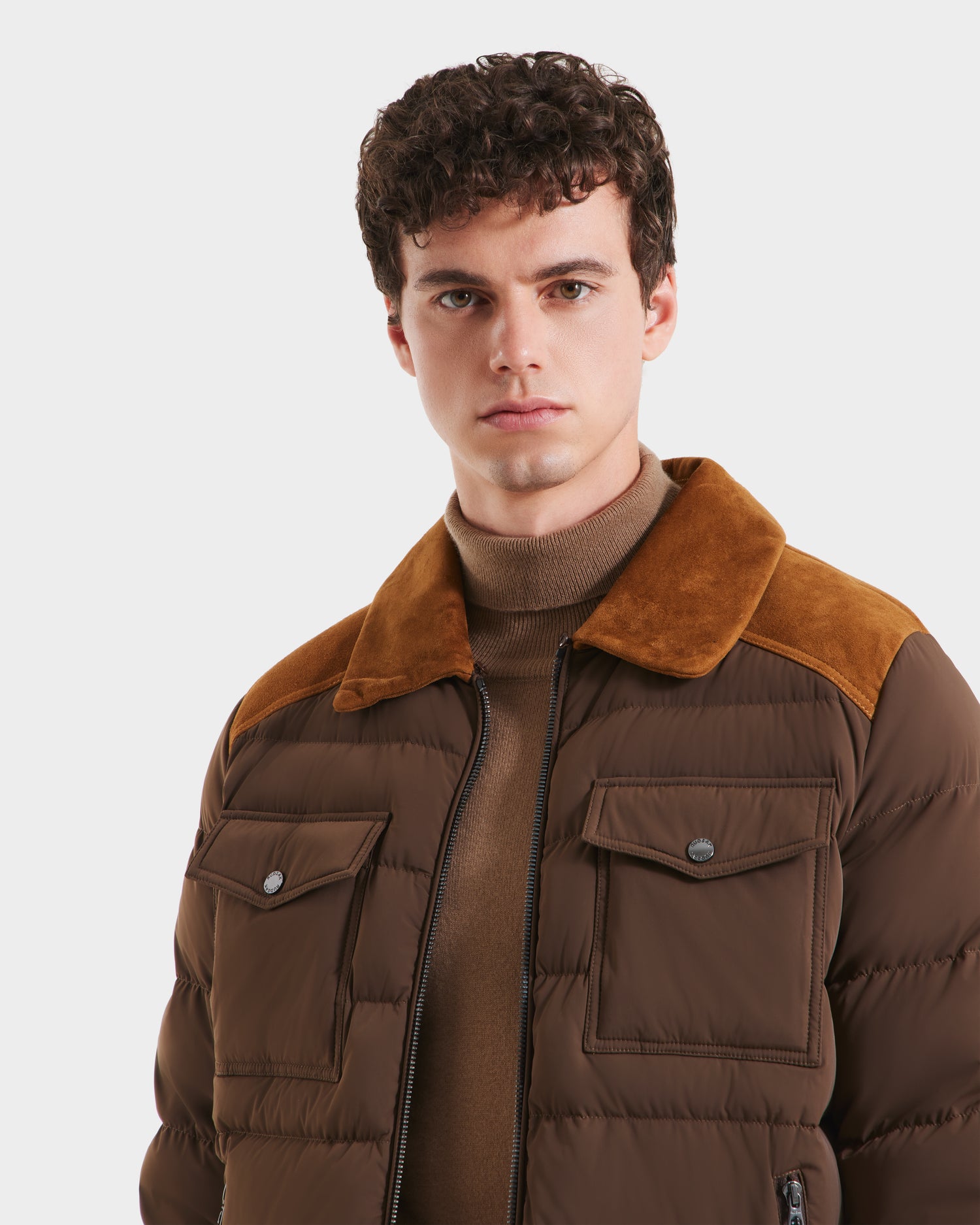 A young man with curly brown hair models the Rudsak ANTHONY - 6125042 Mocha quilted jacket, featuring suede accents and a camel turtleneck, set against a plain white background.