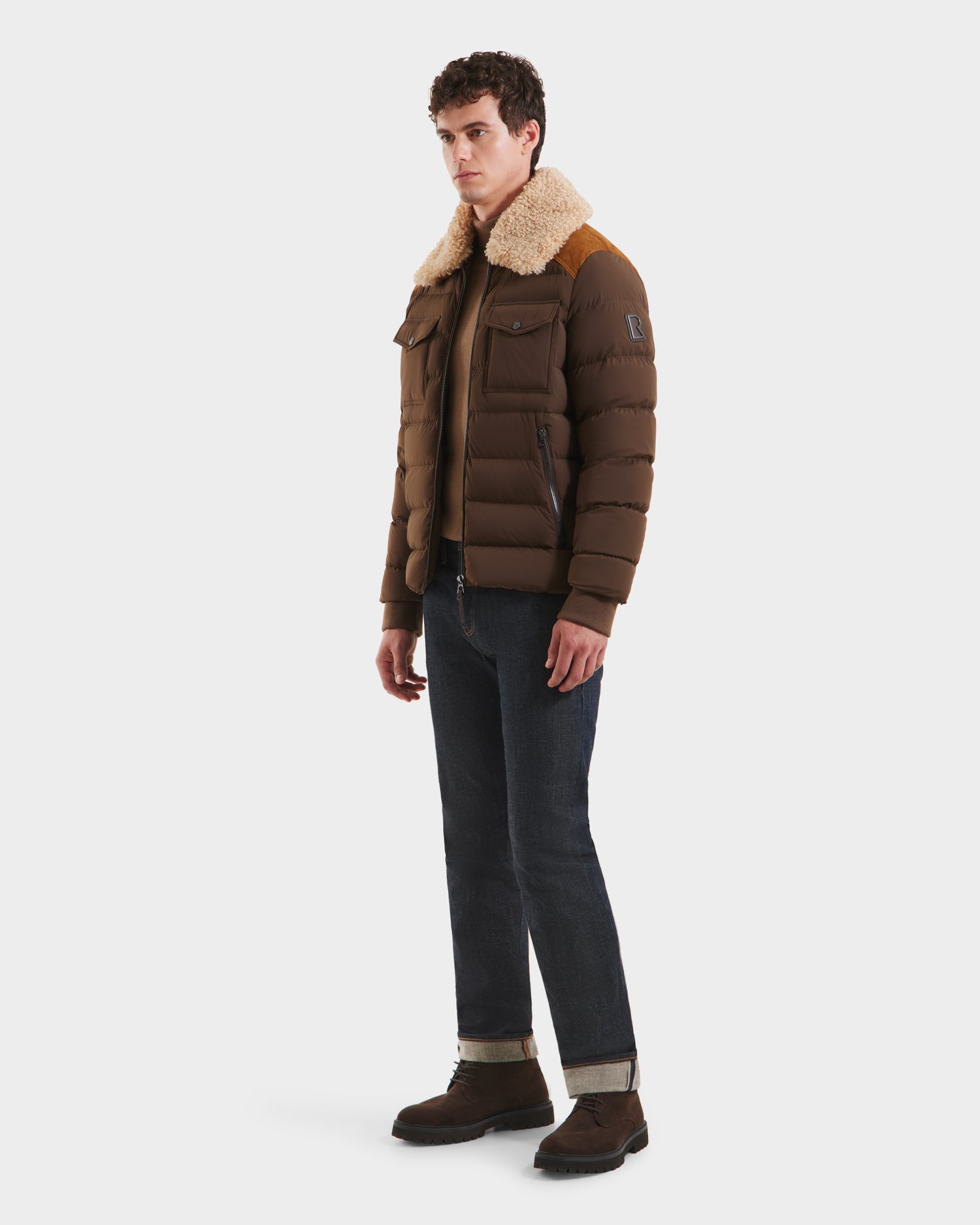A man stands sideways against a plain white background, wearing Rudsak’s ANTHONY - 6125042 Mocha quilted jacket with a shearling collar, paired with dark jeans and brown shoes.