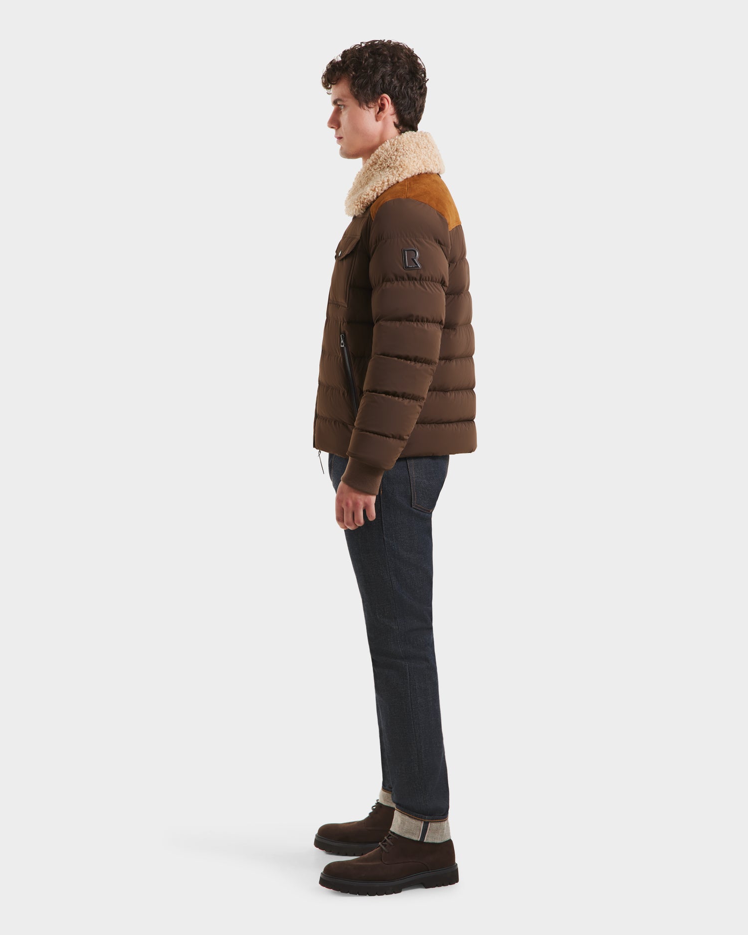 A man stands in profile facing left, wearing the Rudsak ANTHONY - 6125042 Mocha men’s aviator jacket with a shearling collar, dark jeans, and dark brown boots against a plain white background.