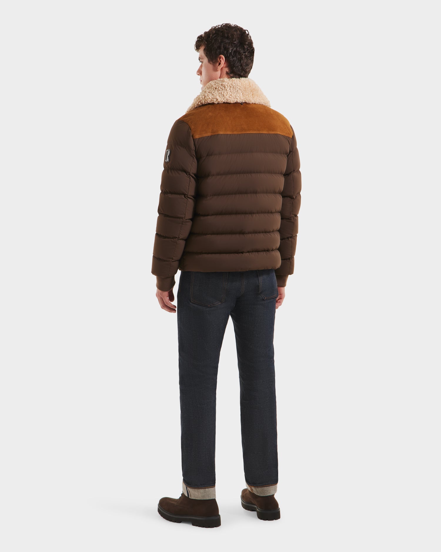 A man faces away against a white background, wearing Rudsaks ANTHONY - 6125042 Mocha brown quilted jacket with a tan shearling collar, paired with dark jeans and brown boots.