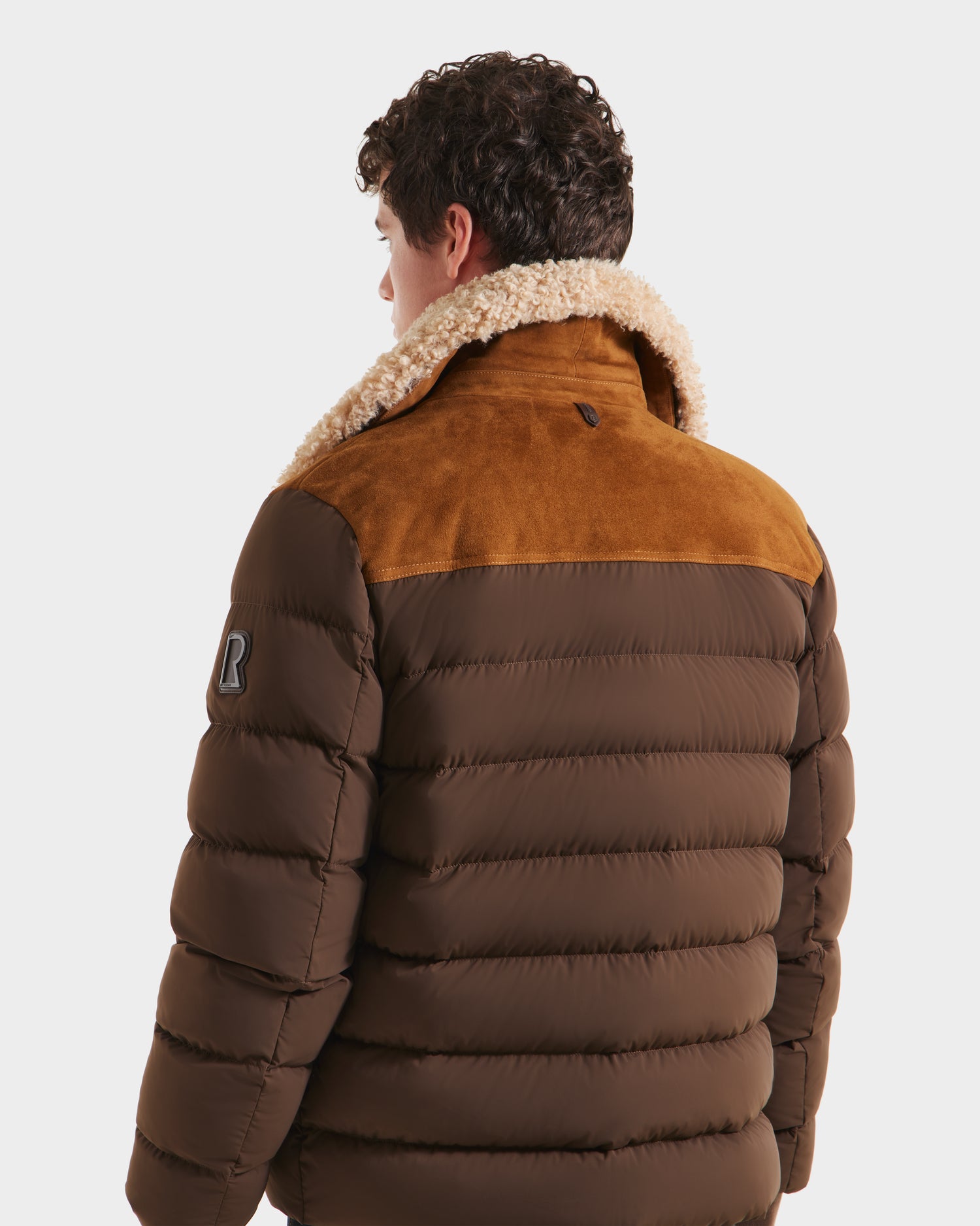 A person with curly hair is shown from the back wearing the Rudsak ANTHONY - 6125042 Mocha men’s aviator jacket, featuring a brown quilted body, tan suede yoke, shearling collar, and an R patch on the sleeve against a white background.