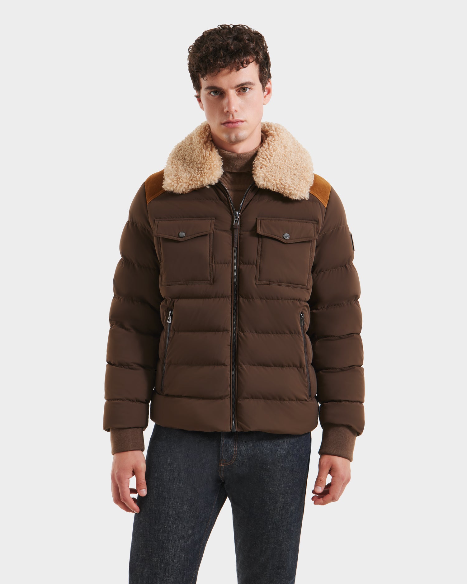 A young man wearing the Rudsak ANTHONY - 6125042 Mocha mens aviator jacket with a light tan shearling collar, front pockets, and zipper closure, paired with dark jeans, stands against a plain white background.
