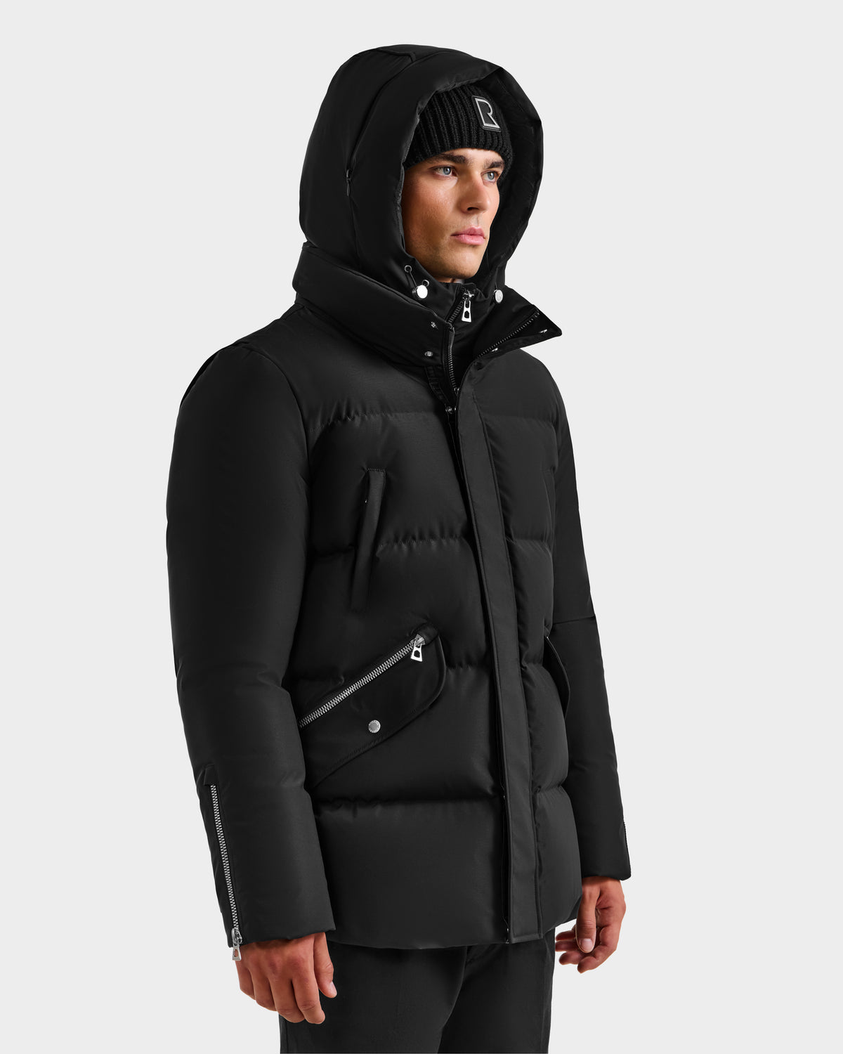 A man wearing a black Rudsak APOLLO H - 6125690 men’s down parka with hood, black beanie, and black pants stands slightly turned against a plain light gray background.