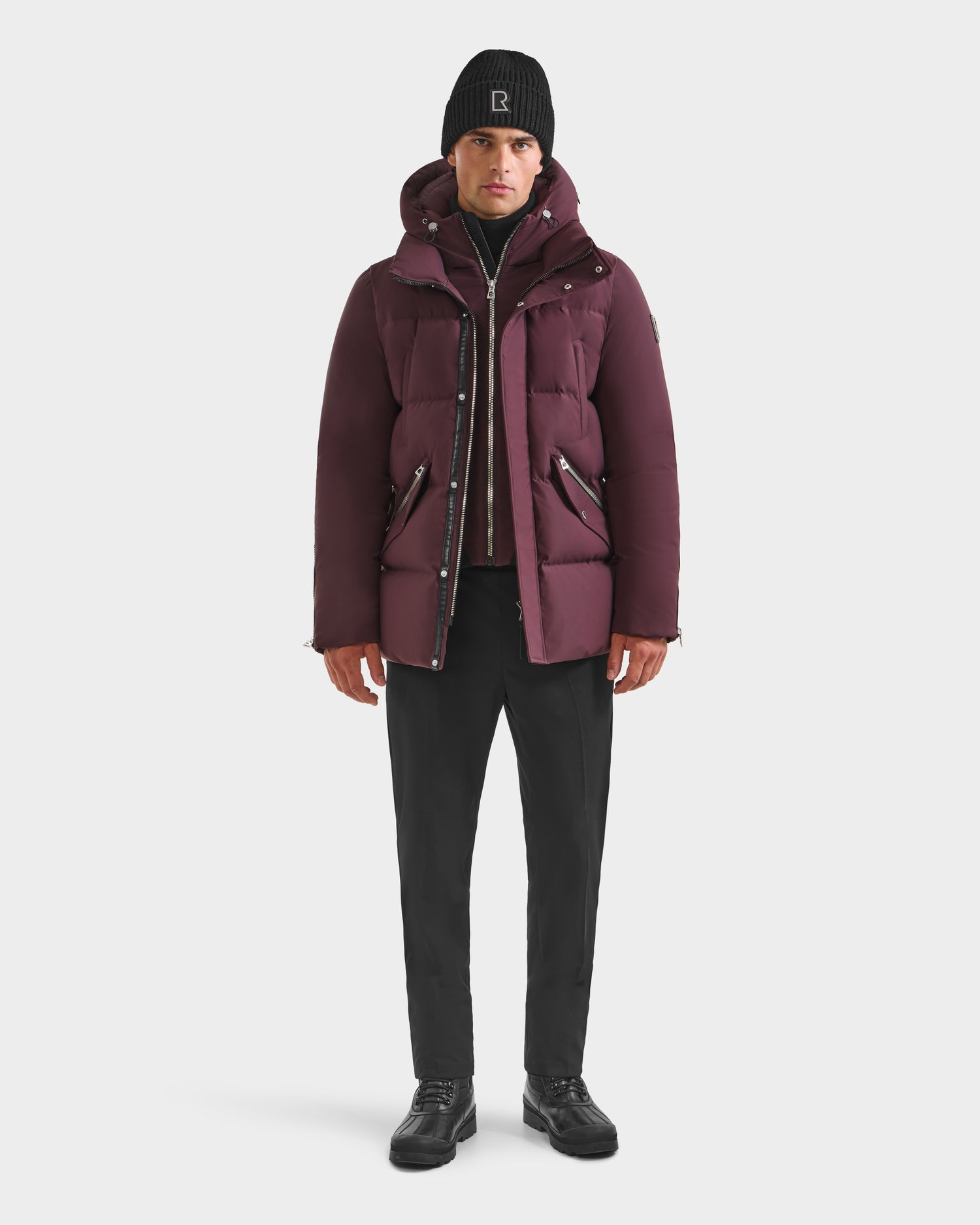 A man wearing the Rudsak APOLLO H - 6125690 Bordeaux down parka, black pants, black shoes, and a black knit beanie stands facing forward against a plain white background.