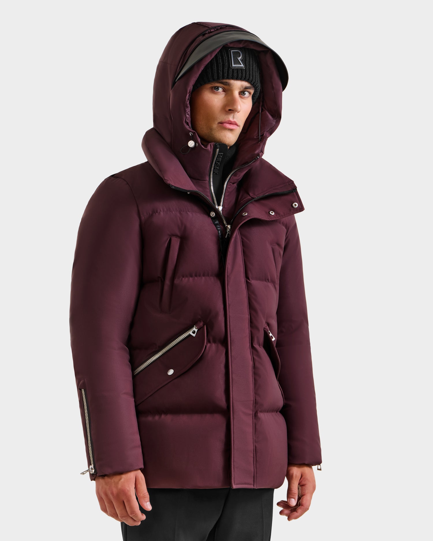 A person in a Rudsak APOLLO H - 6125690 Bordeaux parka with zippered pockets and high-loft down fill, paired with a black beanie, stands against a plain light gray background.