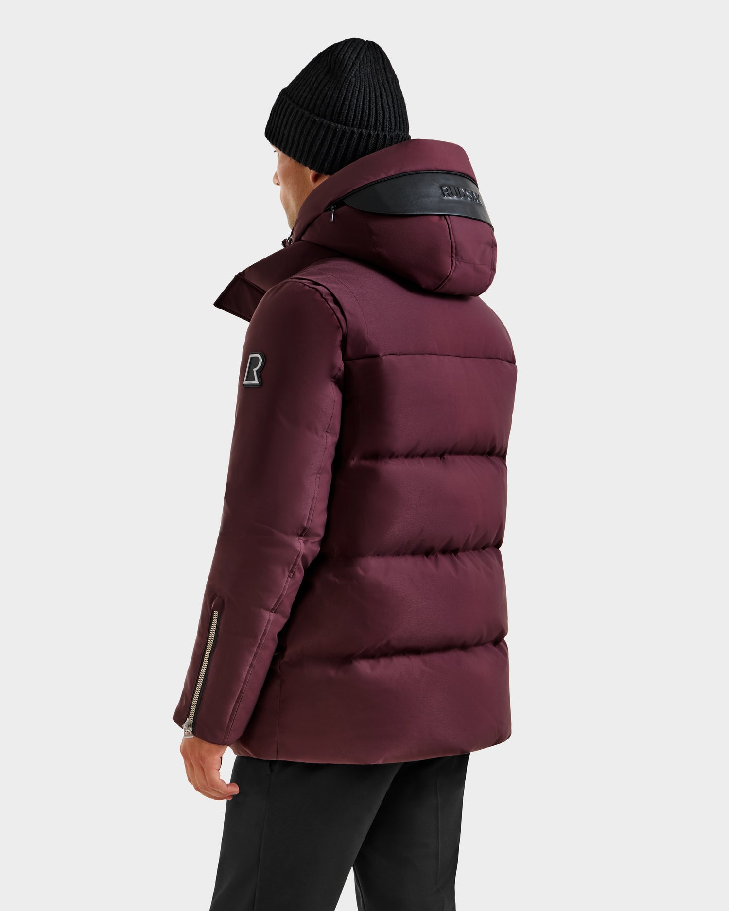 A person wearing the Rudsak APOLLO H - 6125690 Bordeaux maroon quilted parka with hood, featuring a black patch on the sleeve, black stripe detail on the hood, and a black beanie, stands with their back turned.