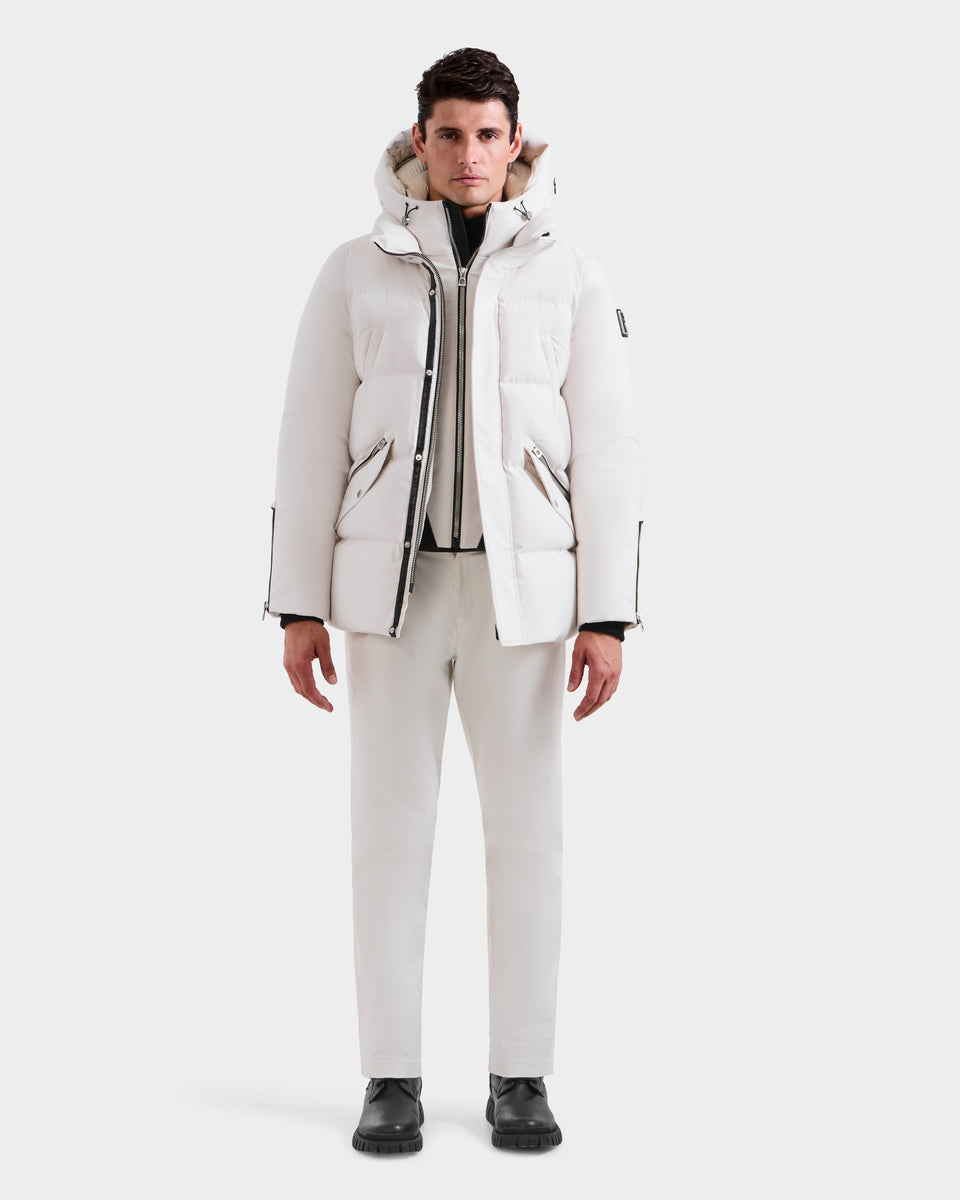 A man stands facing forward in a Rudsak APOLLO H - 6125690 Vanilla parka, with matching white pants and black boots, against a plain white background.