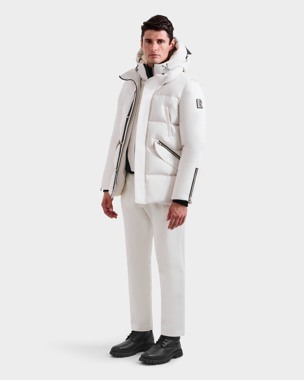 A man wears the Rudsak APOLLO H - 6125690 Vanilla men's quilted down parka, styled with white pants and black boots against a plain white background. The winter parka features zippered pockets and an upper sleeve patch.