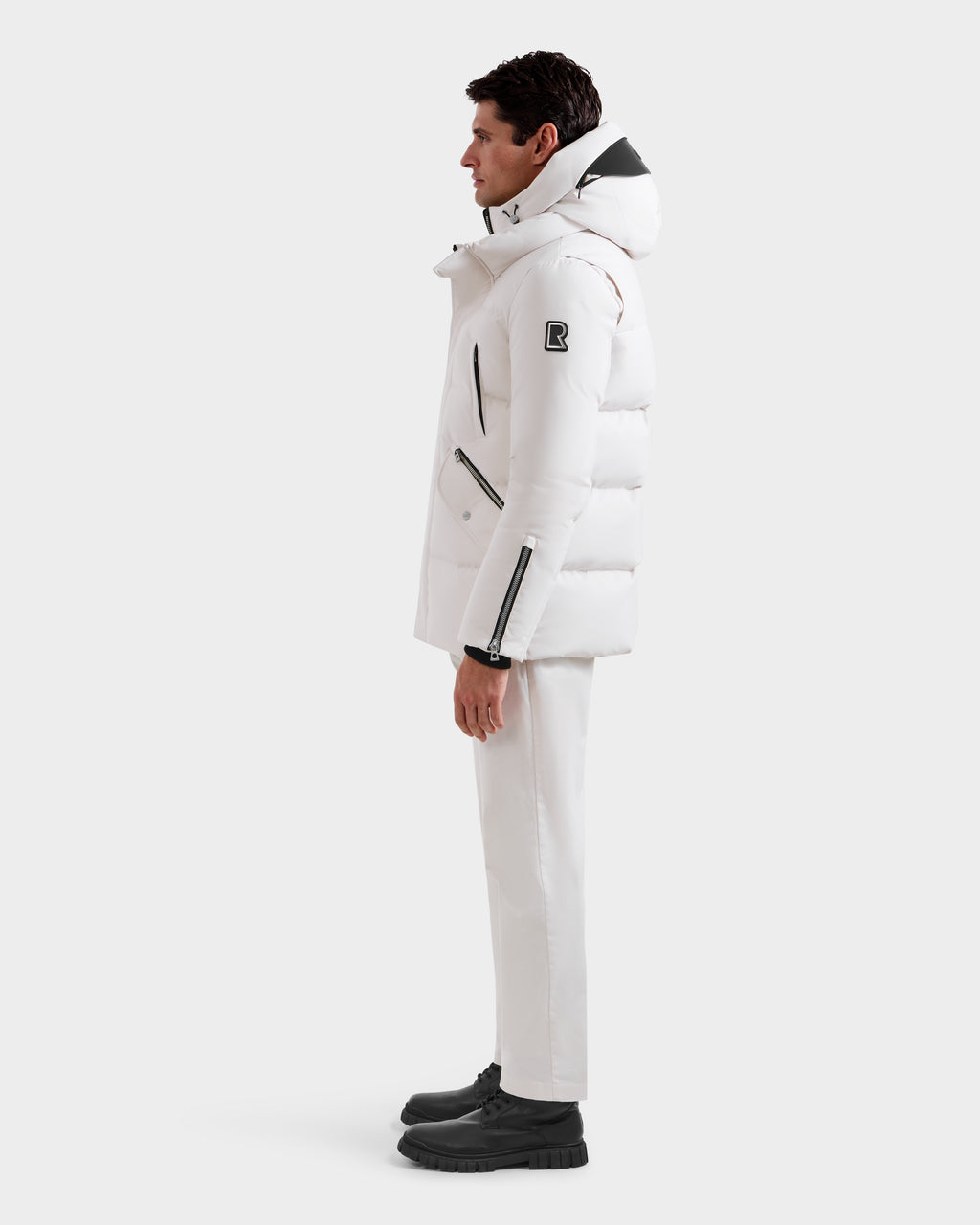 A man in profile wears the Rudsak APOLLO H - 6125690 Vanilla men’s quilted down parka, paired with white pants and black boots. The winter parka features black zipper accents and a distinctive patch on the upper sleeve, set against a white background.