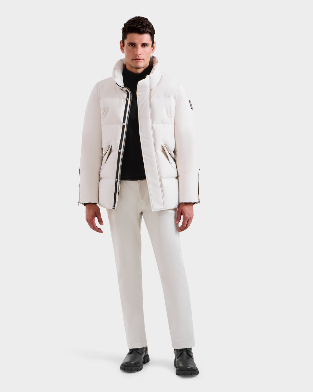 A man stands facing forward, wearing the Rudsak APOLLO H - 6125690 Vanilla men’s quilted down parka, a black turtleneck sweater, white pants, and black shoes against a plain white background.