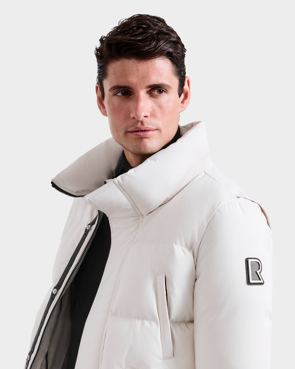 A man with short dark hair wears the Rudsak APOLLO H - 6125690 Vanilla winter parka, featuring a white quilted design, high collar, and black R patch on the sleeve, as he looks slightly to his left against a plain light background.